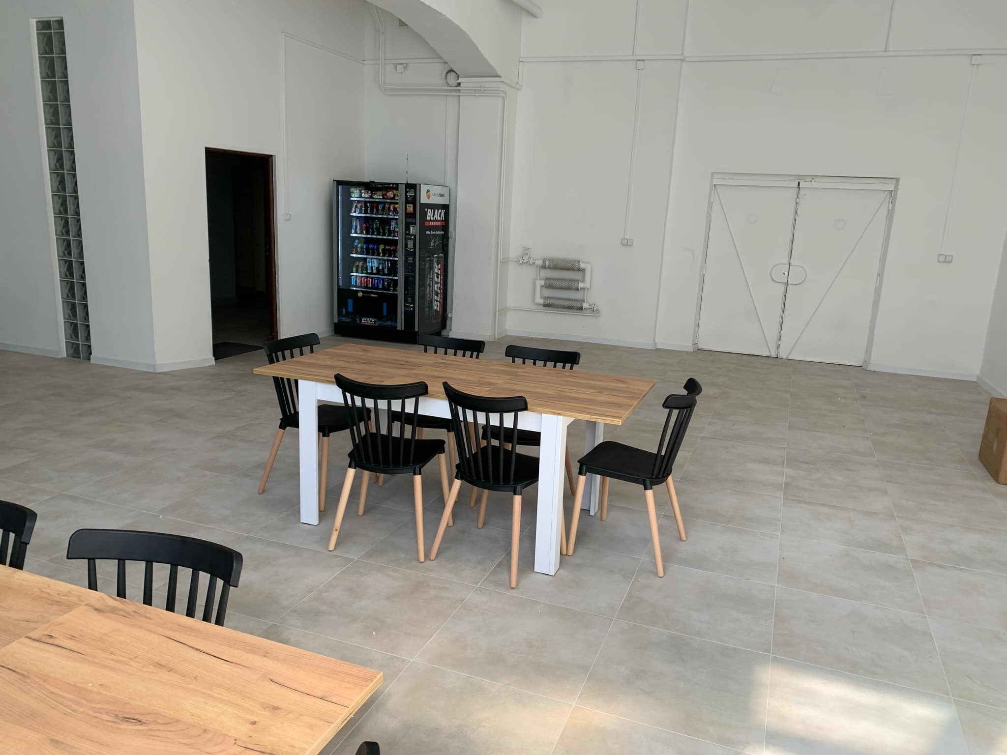 Jasna, przestronna sala z płytkami podłogowymi, stołem z krzesłami i automatem z napojami. Minimalistyczny wystrój, neutralna kolorystyka.
