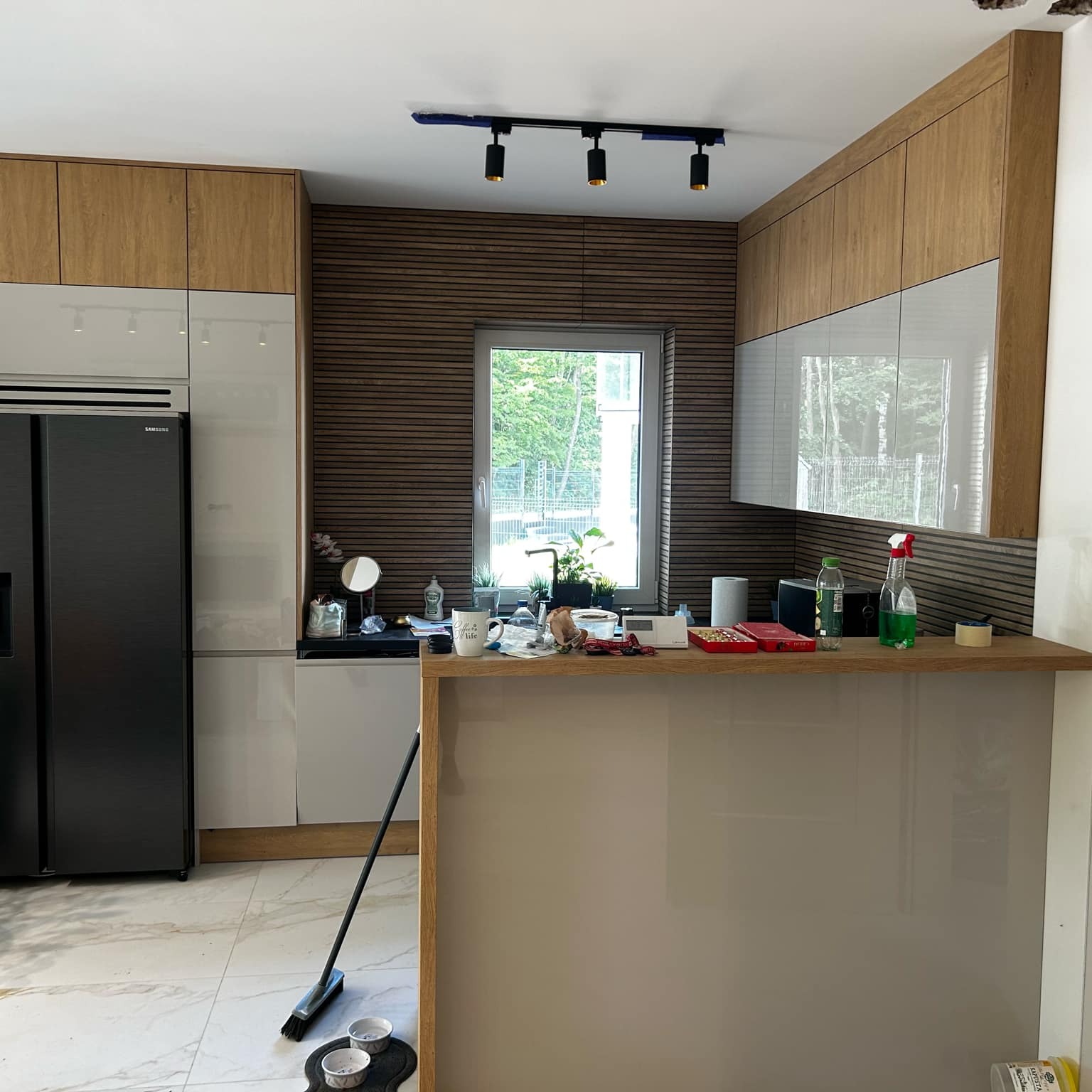 Nowoczesna kuchnia po remoncie: biało-drewniane szafki, czarna lodówka, marmurowe płytki. Widoczne detale wykończenia i oświetlenie punktowe.