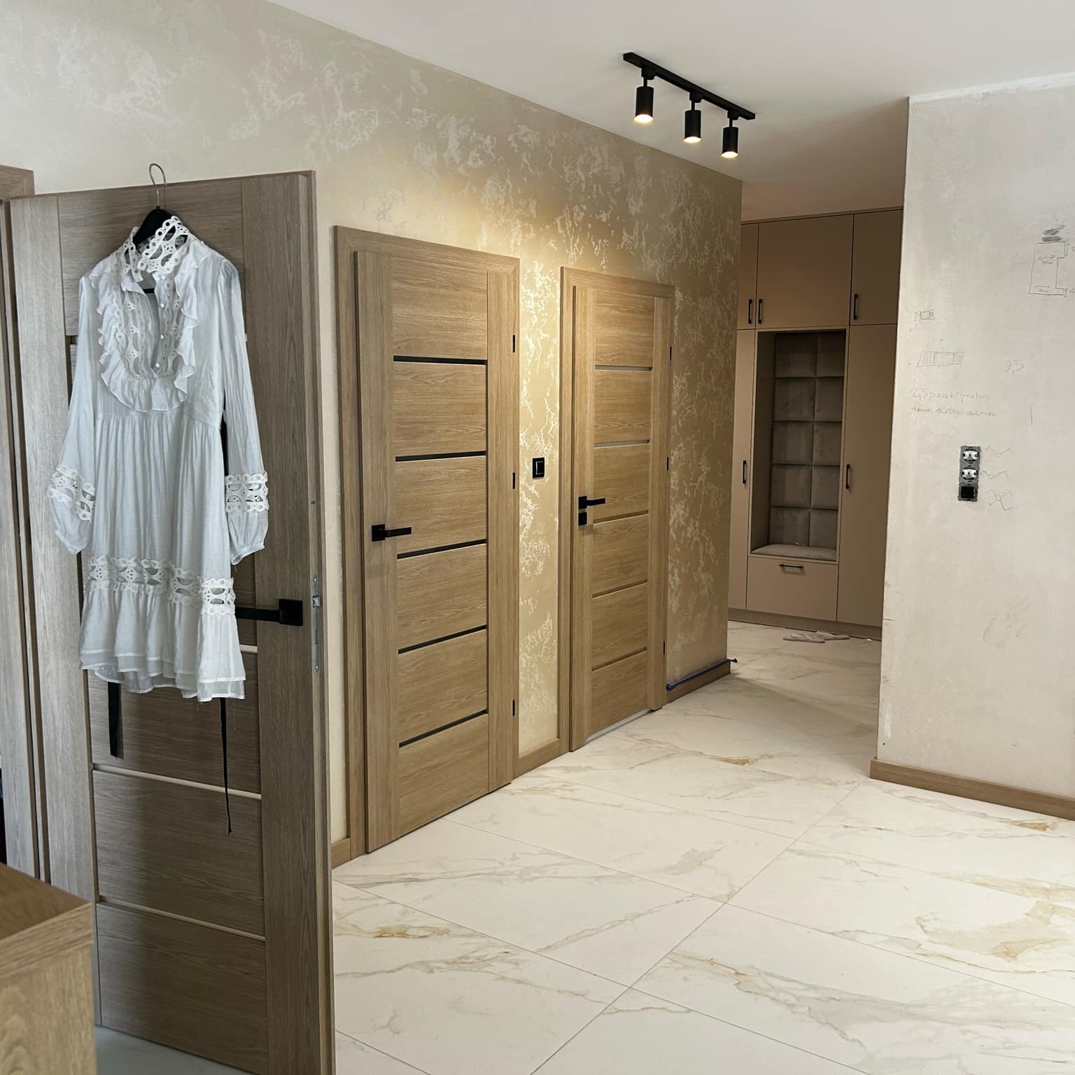 Wykończone wnętrze: jasne płytki podłogowe imitujące marmur, drewniane drzwi wewnętrzne z czarnymi klamkami, beżowa ściana z dekoracyjną fakturą i garderoba w tle.