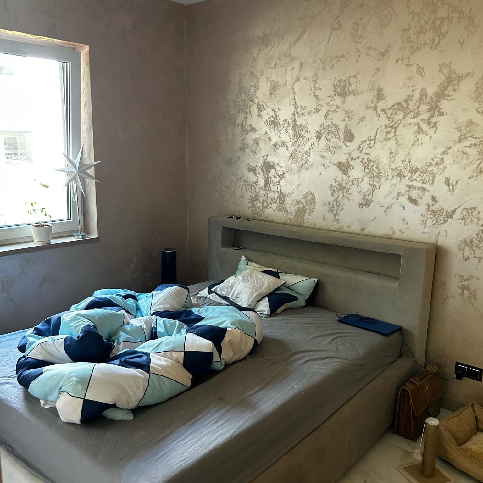 Wykończenie sypialni: ściana z dekoracyjnym tynkiem w odcieniach beżu i złota, łóżko z szarą ramą i kolorową pościelą, okno z ozdobą. Nowoczesny design.