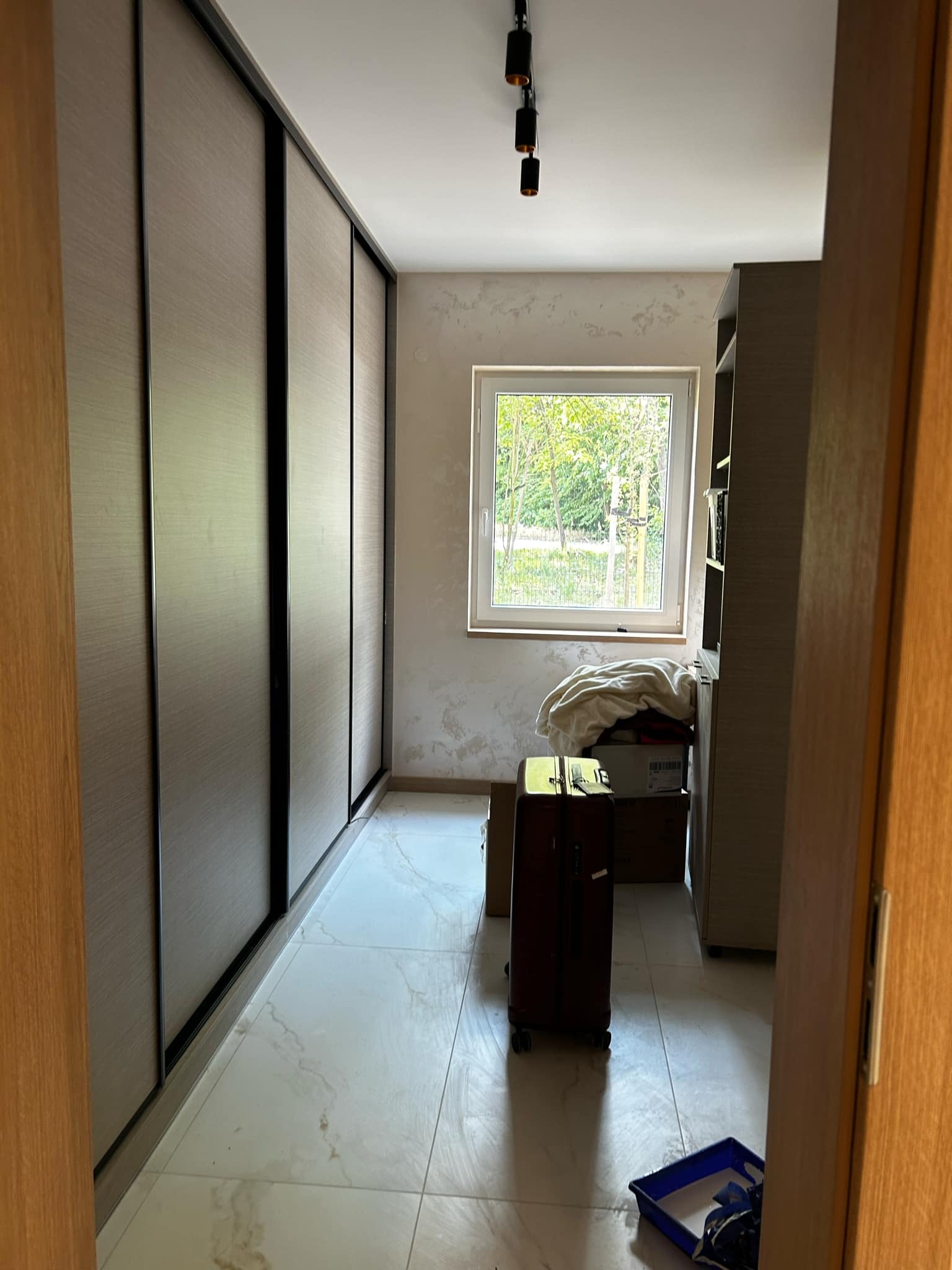 Wnętrze pokoju z dużą szafą przesuwną z drewnianymi frontami, marmurową podłogą i oknem. Widoczny bagaż i akcesoria malarskie.