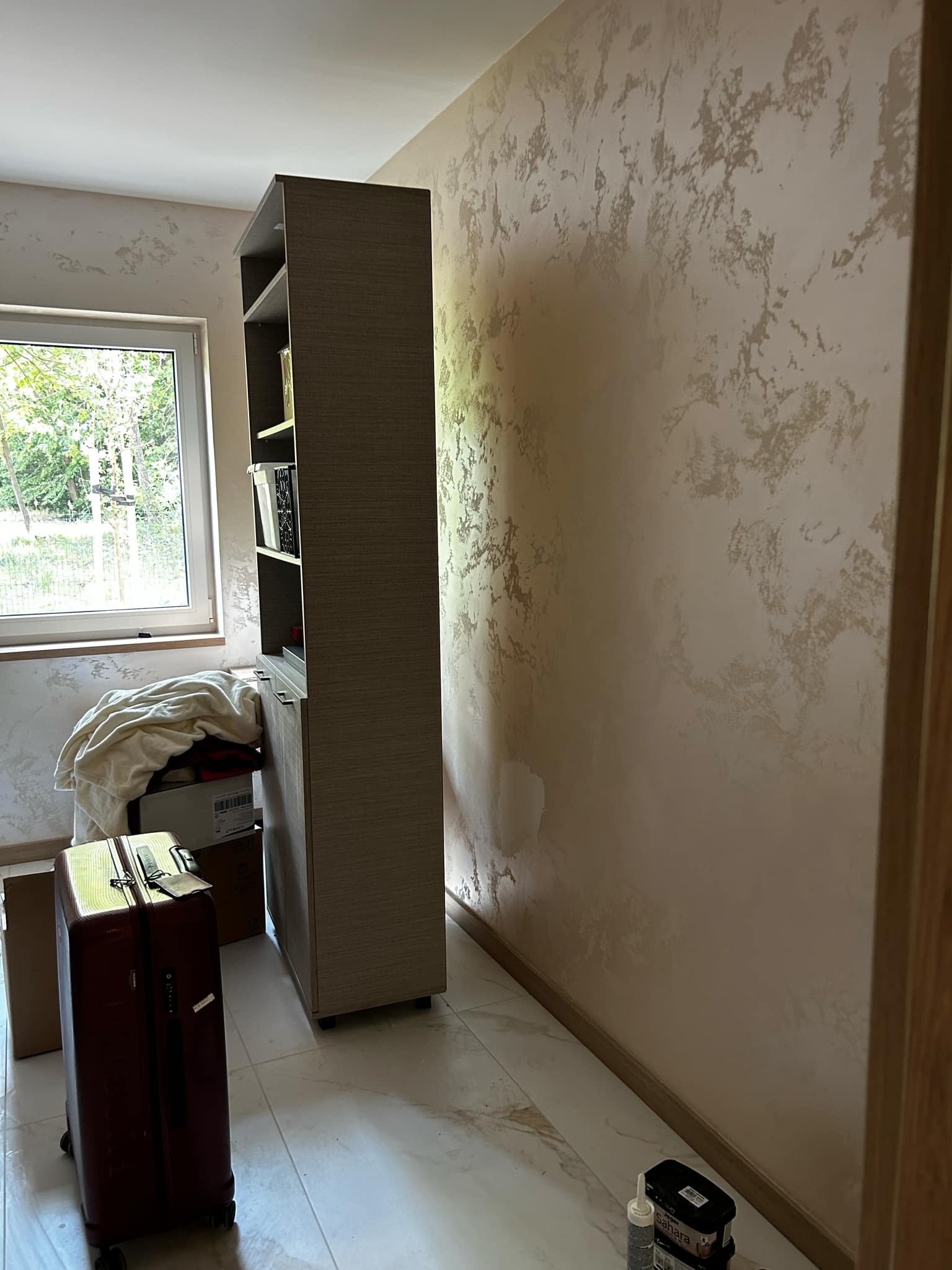 Ściana z efektem dekoracyjnym w Częstochowie, widoczna obok regału i okna. Na podłodze leżą materiały budowlane i walizka, co wskazuje na trwający remont.