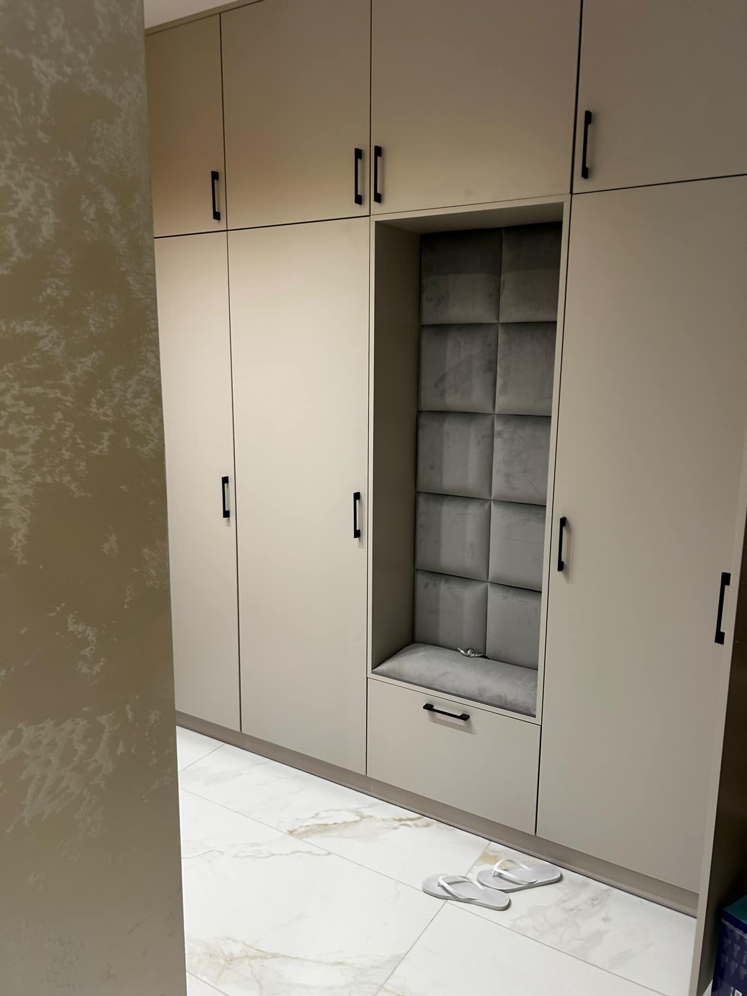 Beżowa zabudowa szafy z siedziskiem wykończonym szarą tapicerką. Na podłodze białe klapki. Ściana z efektem dekoracyjnym w odcieniach beżu. Nowoczesny design.