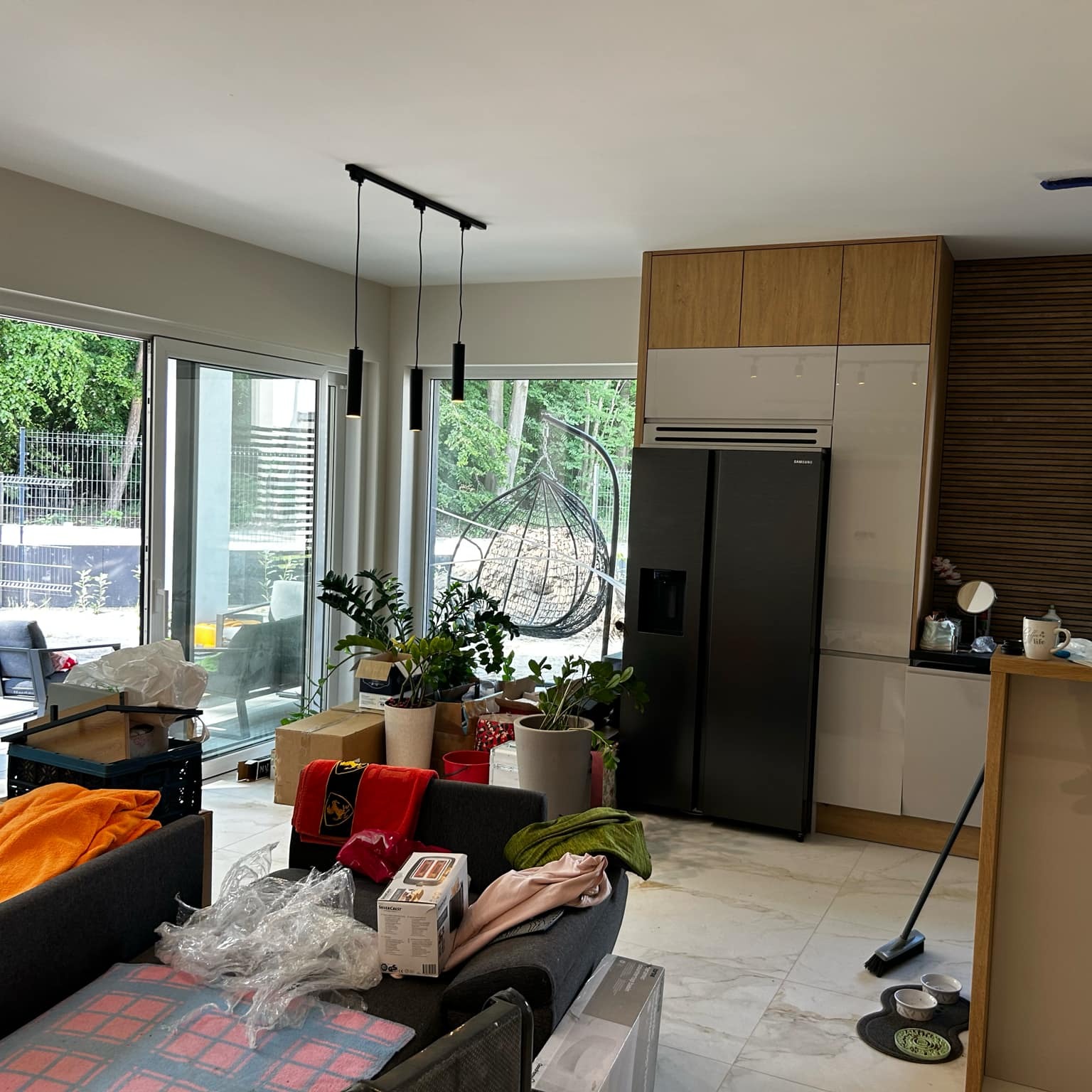 Wnętrze domu w trakcie przeprowadzki lub remontu. Widoczne meble kuchenne, sprzęt AGD, kartony i inne przedmioty. Duże okna z widokiem na zieleń.