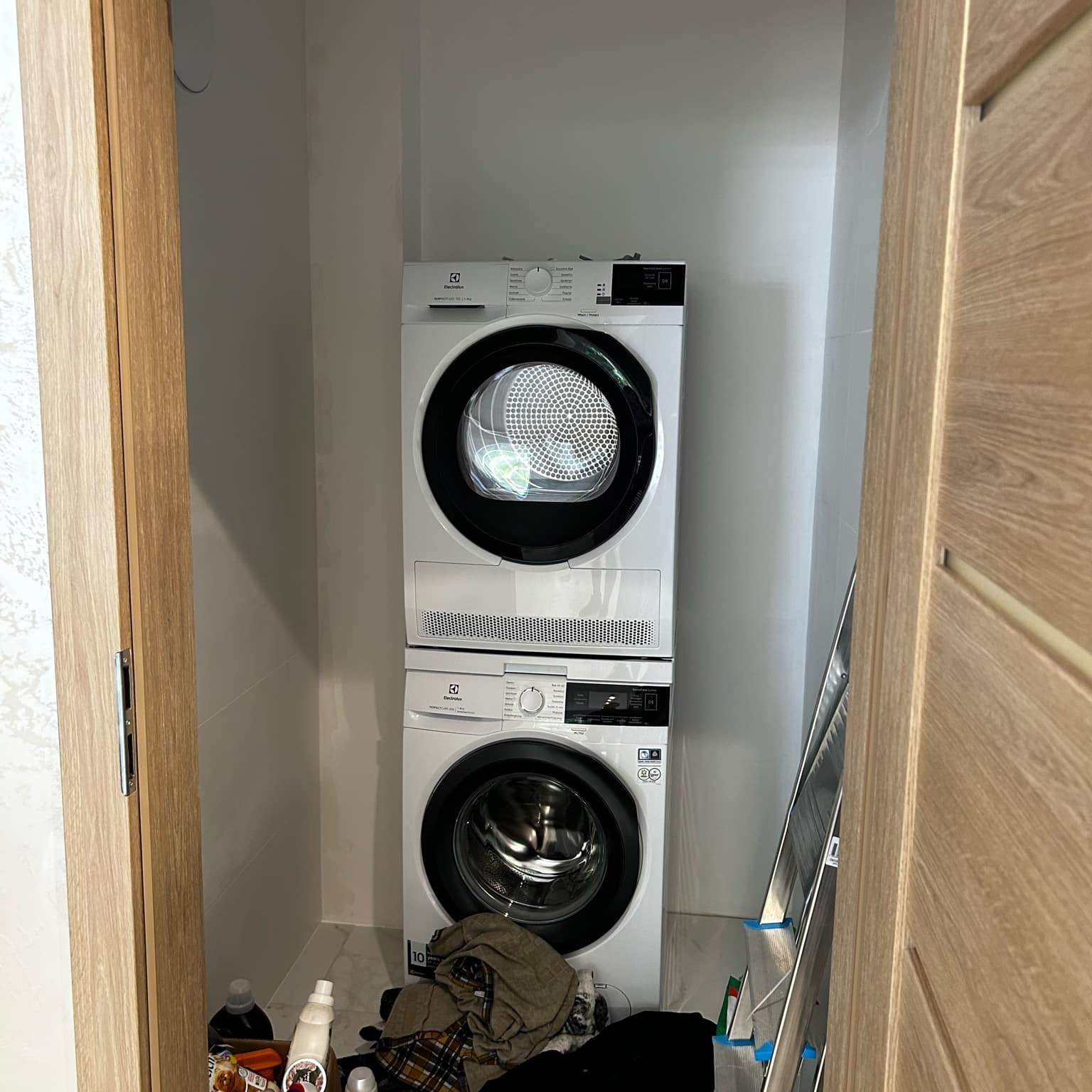 Ustawiona pralka i suszarka Electrolux w jasnej łazience, widok zza otwartych drzwi. W tle białe płytki i drabina. Na podłodze środki czystości.