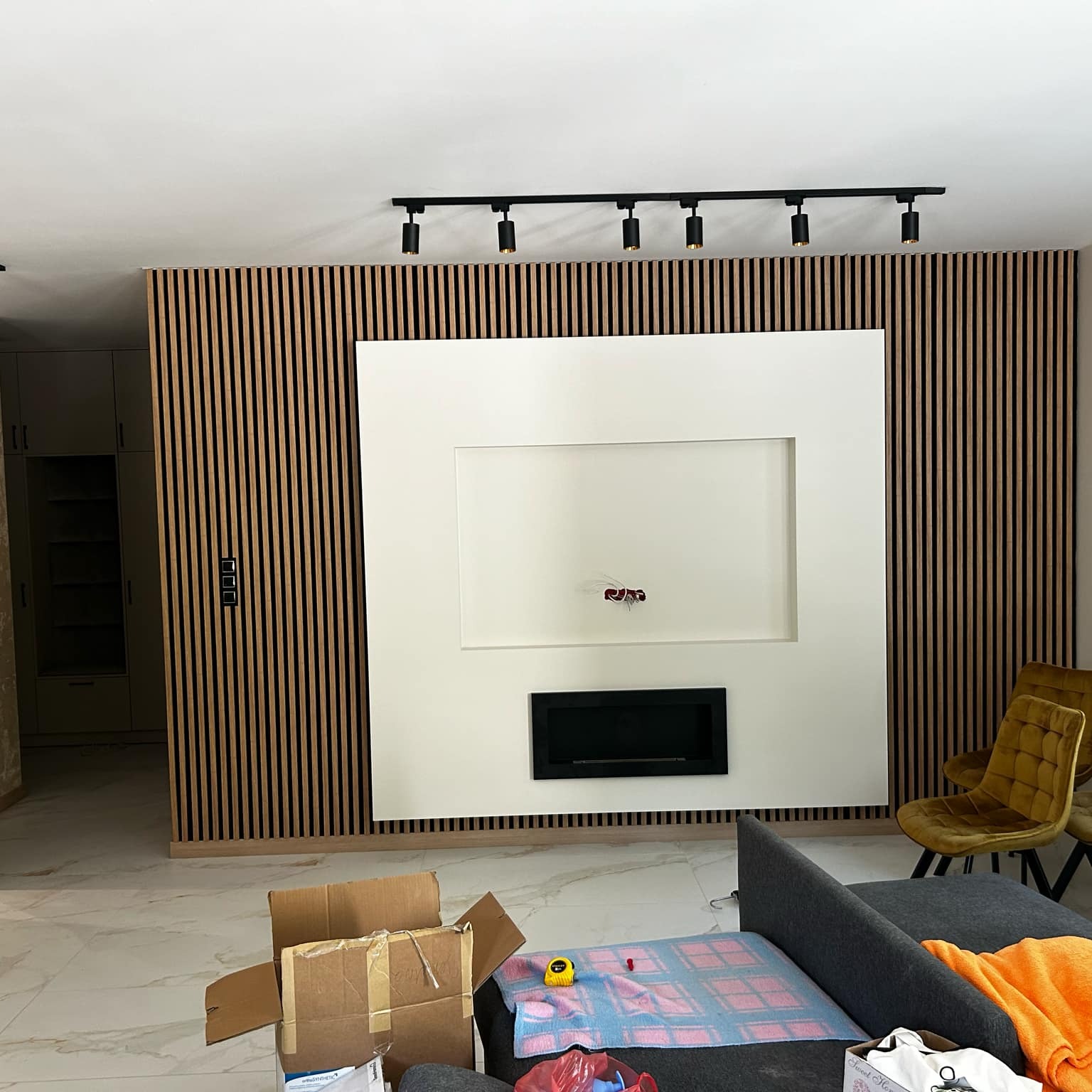 Wykończona ściana z drewnianych lameli z wnęką na TV i kominkiem elektrycznym, oświetlenie szynowe sufitowe, widok na marmurową podłogę i meble w salonie.