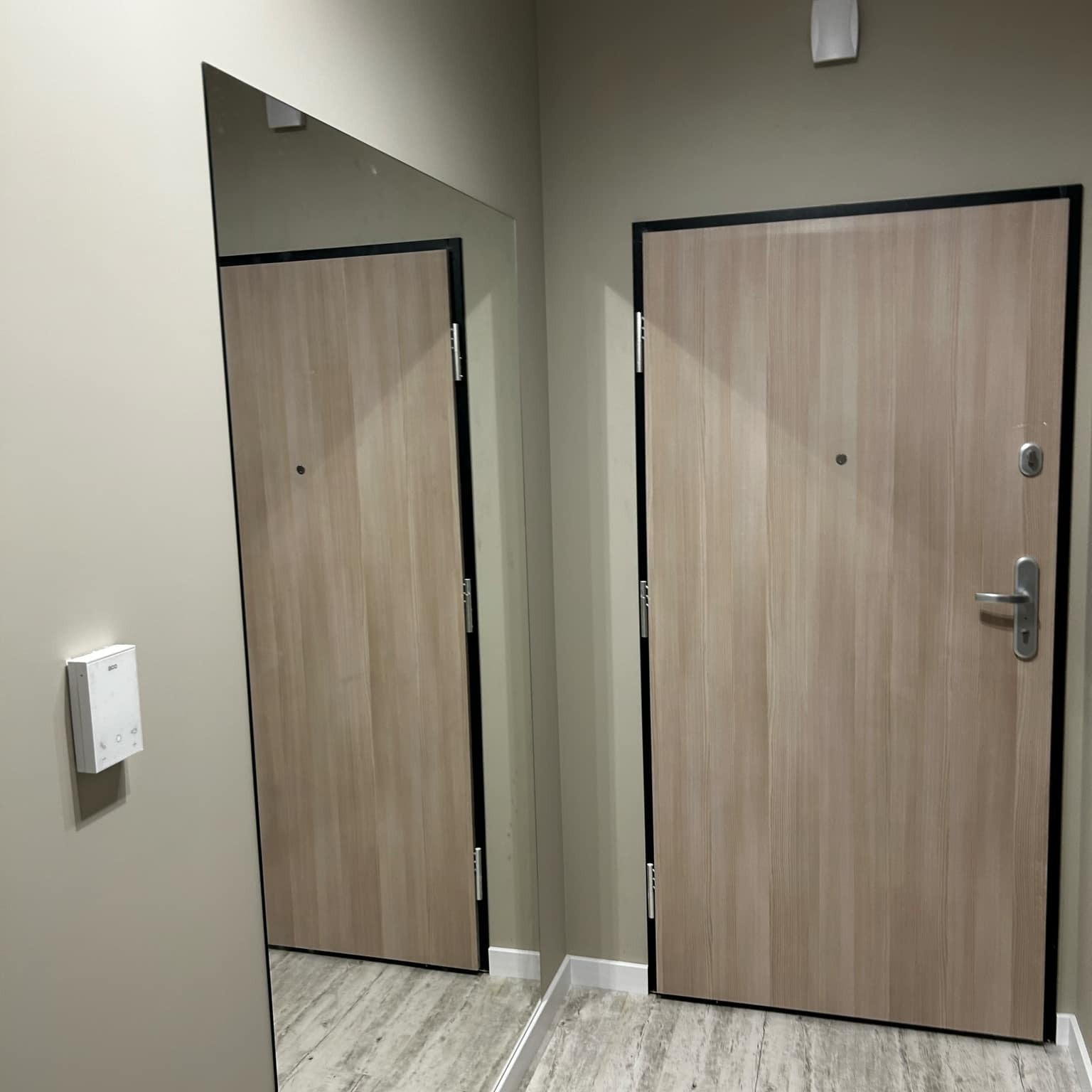 Zamontowane drzwi wejściowe z czarną ramą, widoczne w lustrze. Jasne panele na podłodze. Na ścianie biały panel sterowania.