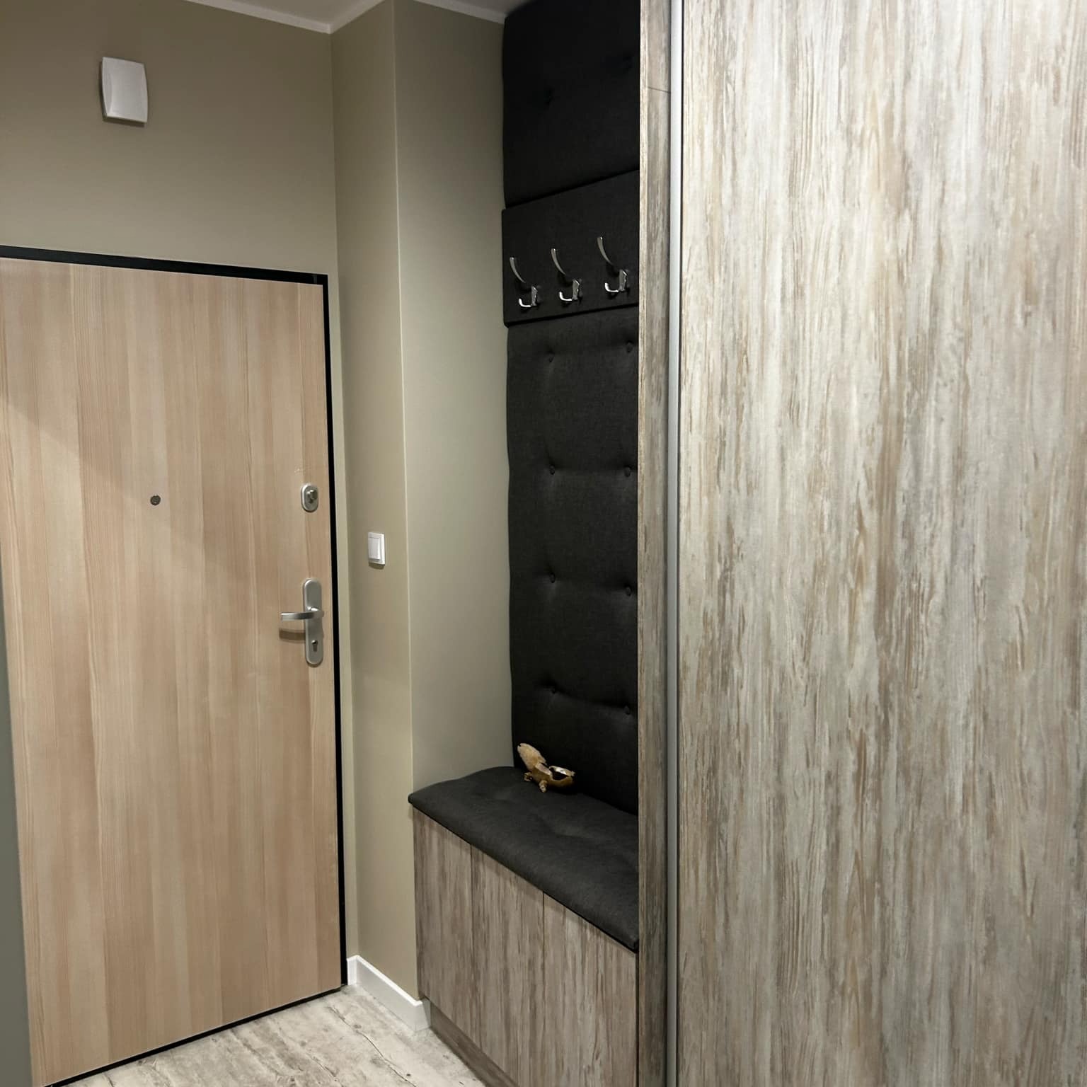 Nowoczesny przedpokój: jasne drewniane drzwi wejściowe, szafa z przesuwnymi drzwiami i tapicerowany panel z wieszakami, tworzący funkcjonalne i stylowe wnętrze.