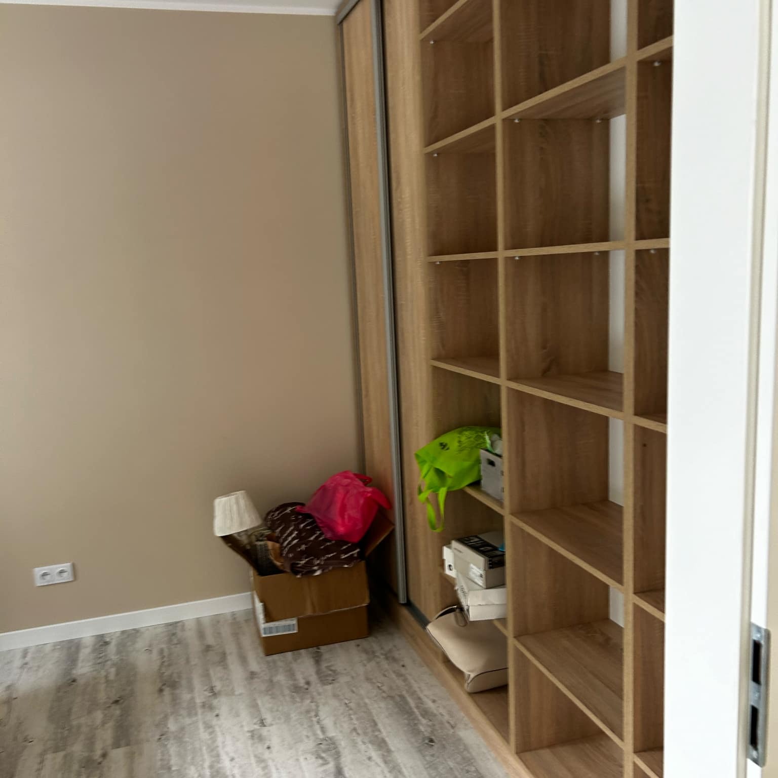 Wnętrze pokoju z beżową ścianą, drewnianą podłogą i zabudową szafy z półkami, częściowo wypełnionymi przedmiotami. W rogu karton z lampą i torbą.