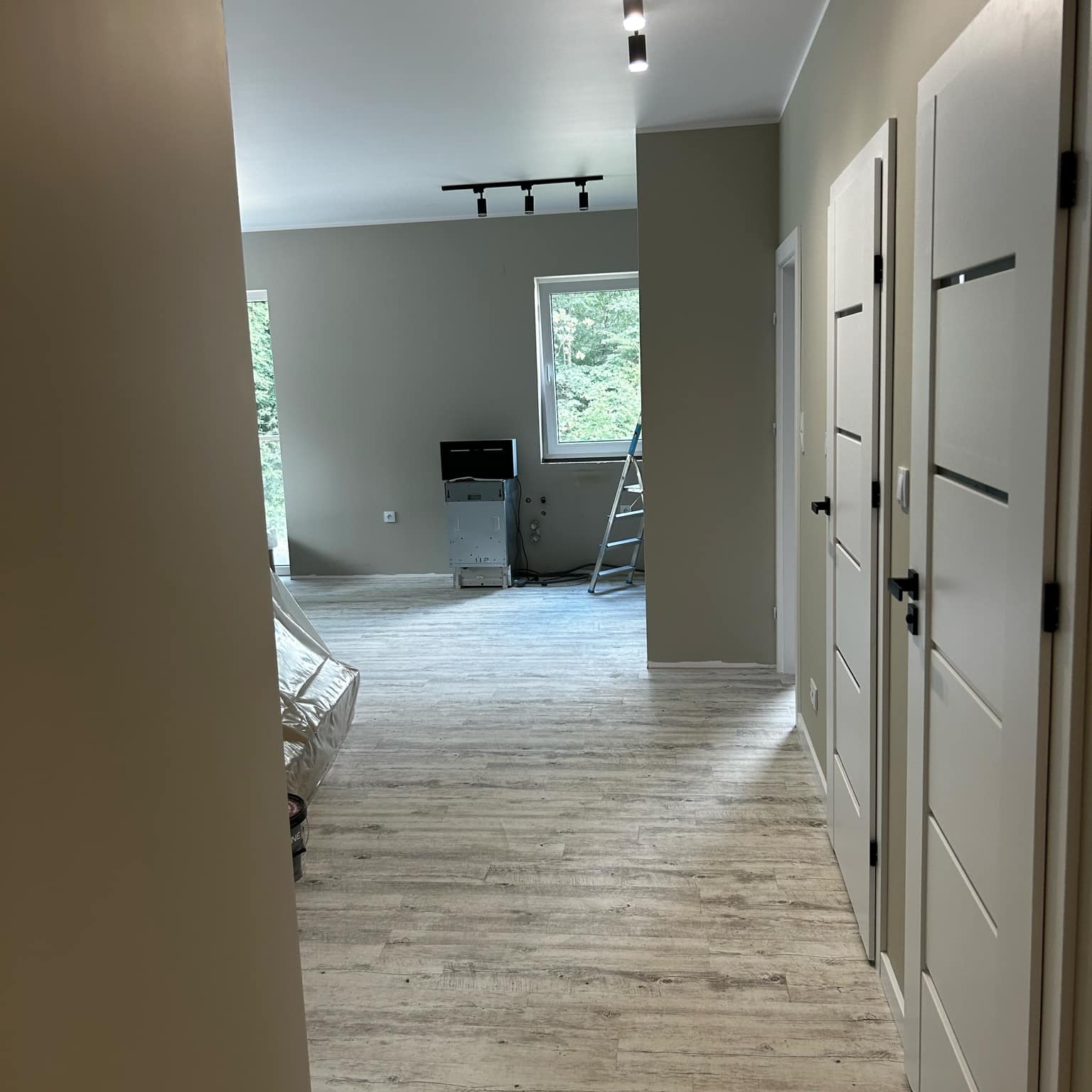 Wnętrze po remoncie: jasne panele podłogowe, świeżo pomalowane ściany w odcieniach szarości, nowe białe drzwi i nowoczesne oświetlenie sufitowe. Widoczna drabina i sprzęt.