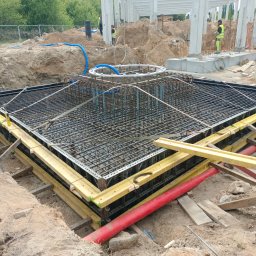 PRO-EKO Paweł Kosiński - Zbrojenie fundamentu pod budynek żelbetowy w wykopie, z widoczną konstrukcją szalunkową i elementami instalacji. Prace ziemne w toku, widoczni pracownicy w tle.