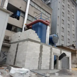 Budowa przyłącza do budynku przemysłowego z białych bloczków. Widoczne rury, komin i elementy instalacji. W tle wysoki budynek z oknami i silosy.