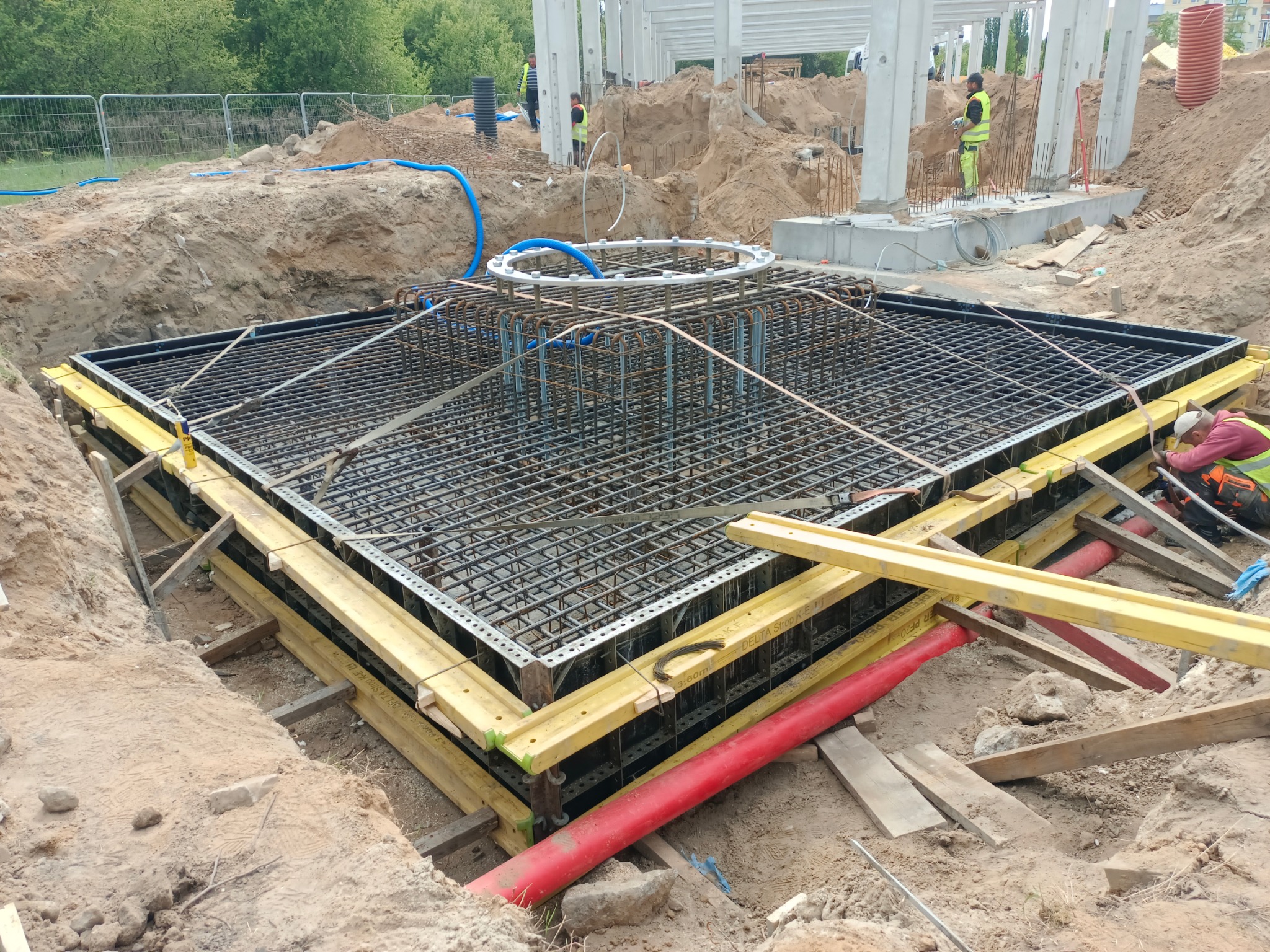Zbrojenie fundamentu pod budynek żelbetowy w wykopie, z widoczną konstrukcją szalunkową i elementami instalacji. Prace ziemne w toku, widoczni pracownicy w tle.