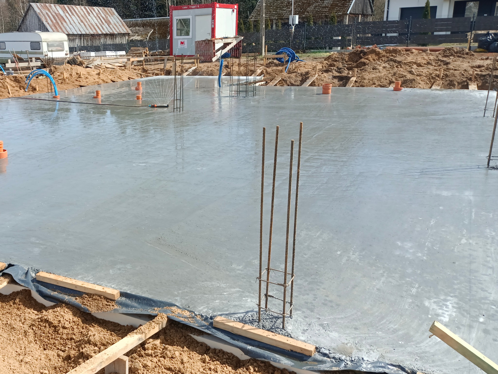 Wylany fundament z wystającymi prętami zbrojeniowymi i rurami instalacyjnymi. Widoczny barak budowlany i teren budowy.