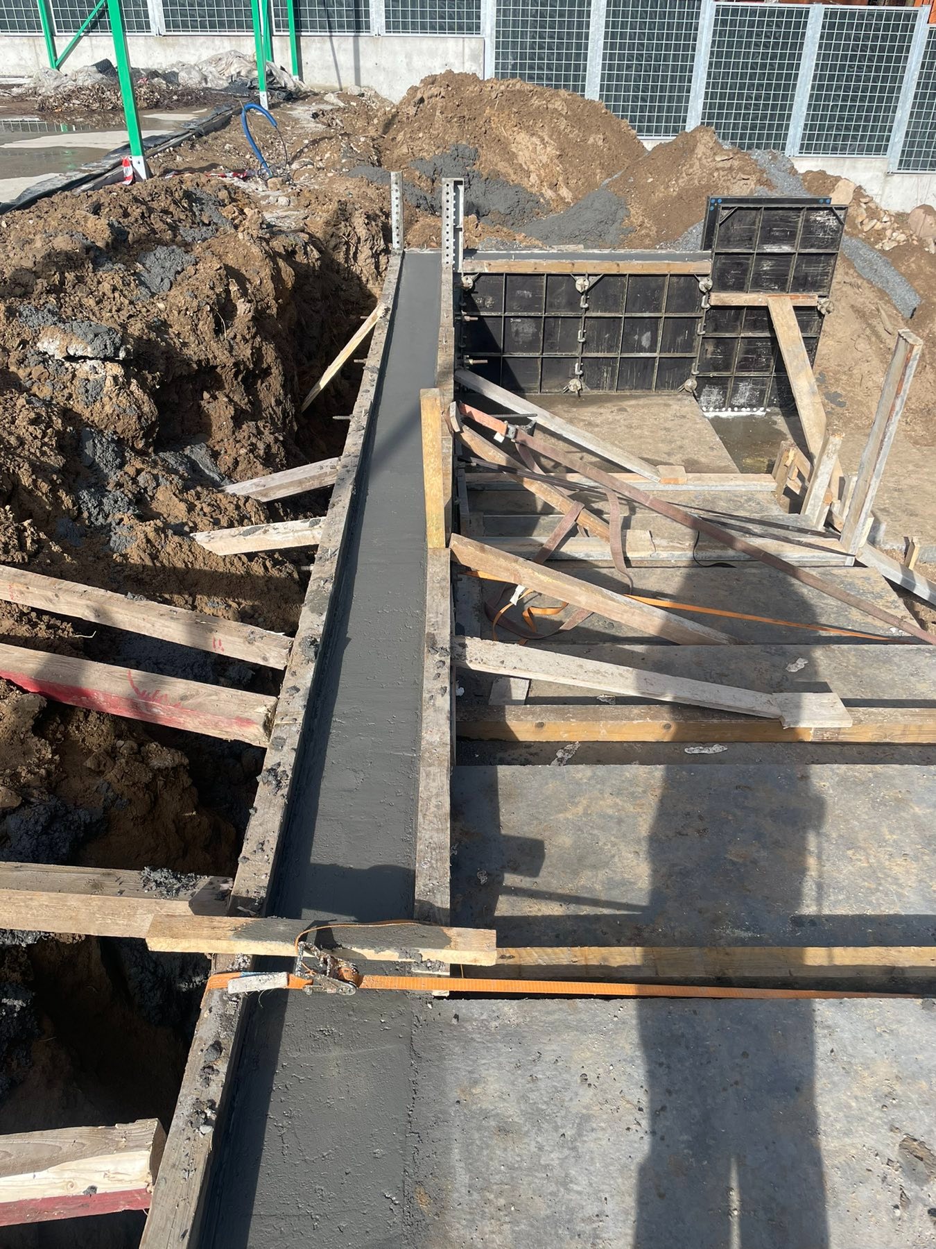 Zalewanie betonem szalunku fundamentu. Widoczne drewniane elementy konstrukcji i świeży beton w szalunku. Ujęcie z góry na placu budowy.