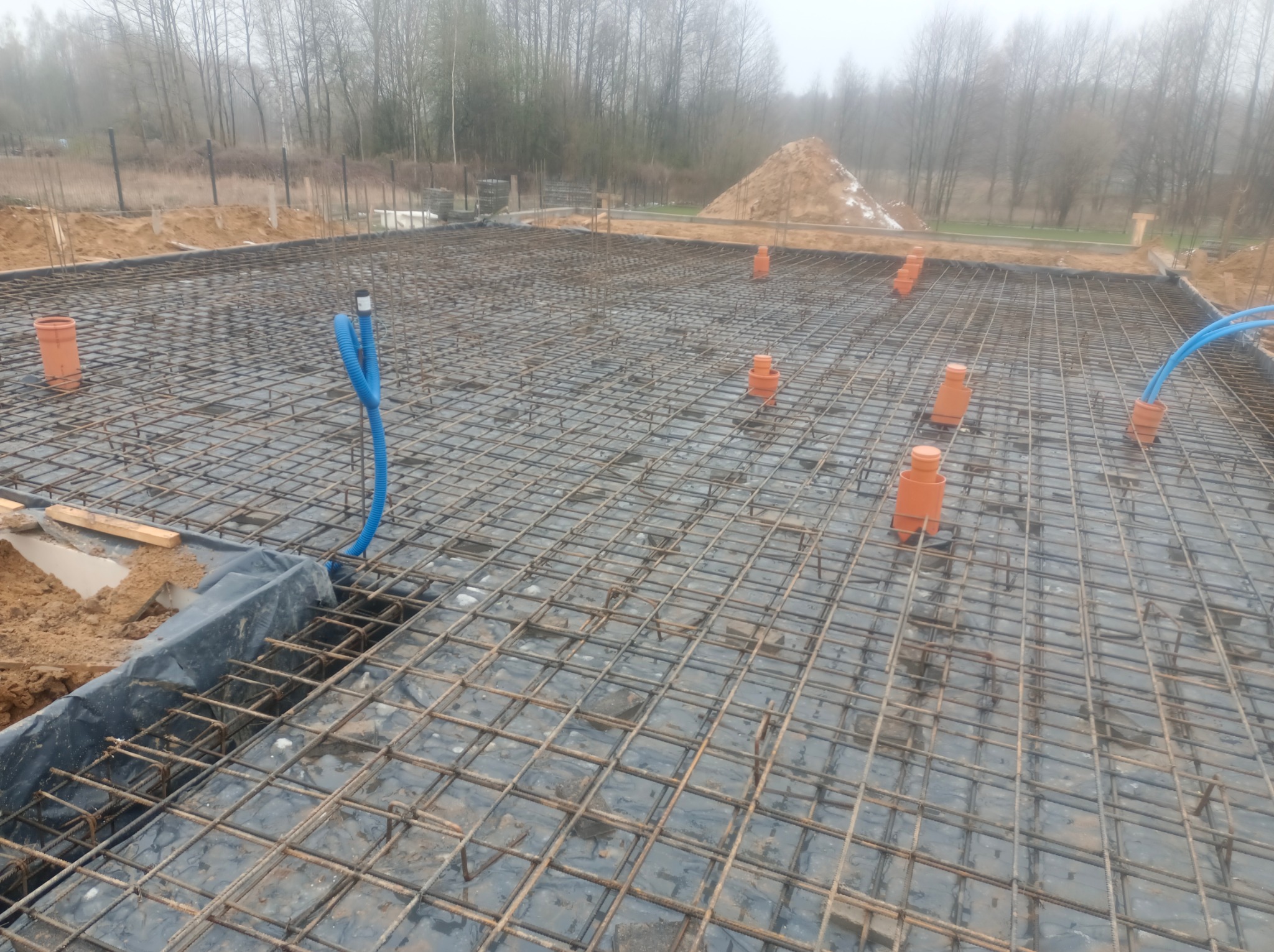 Zbrojenie płyty fundamentowej z wystającymi rurami kanalizacyjnymi i niebieskim peszlem w otoczeniu ziemi i drzew w tle. Widok z góry na przygotowany teren pod budowę.