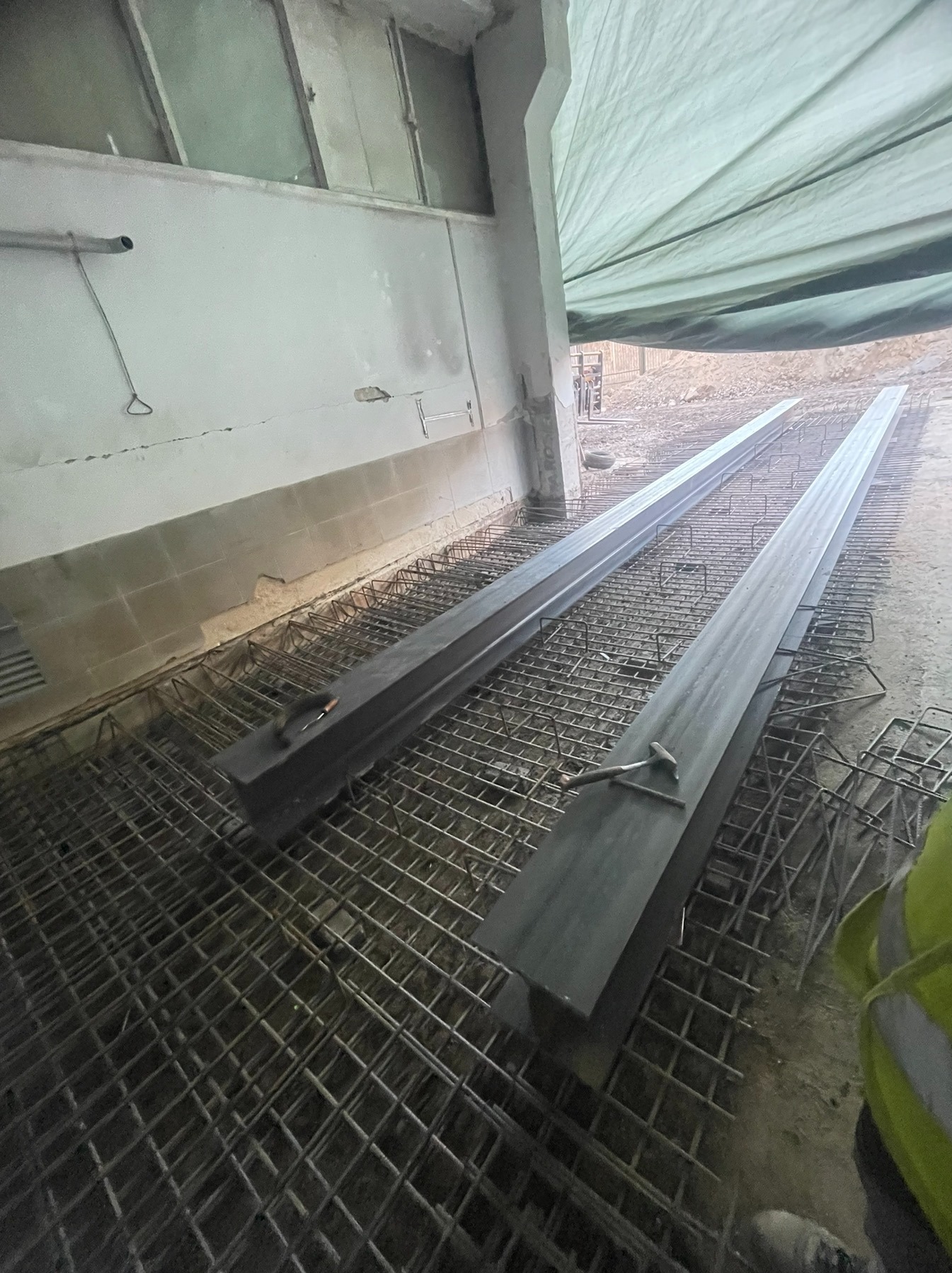 Przygotowanie zbrojenia fundamentów z użyciem metalowych belek i siatki zbrojeniowej. Widoczny fragment narzędzia. Prace budowlane w surowym wnętrzu.