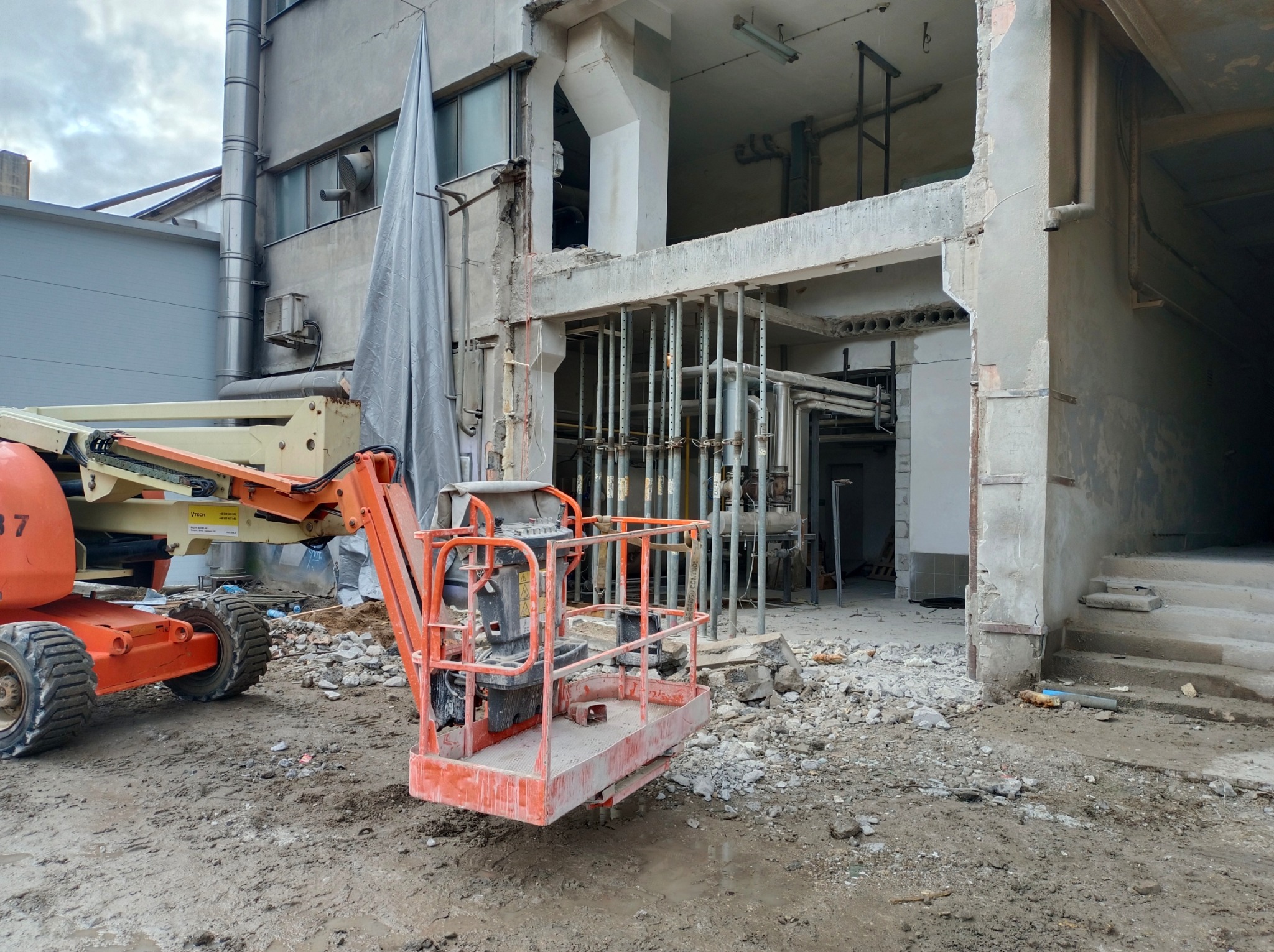 Demontaż konstrukcji żelbetowej. Widoczny podnośnik koszowy i elementy podtrzymujące strop. Gruz na ziemi. W tle fragment budynku z odsłoniętą konstrukcją.