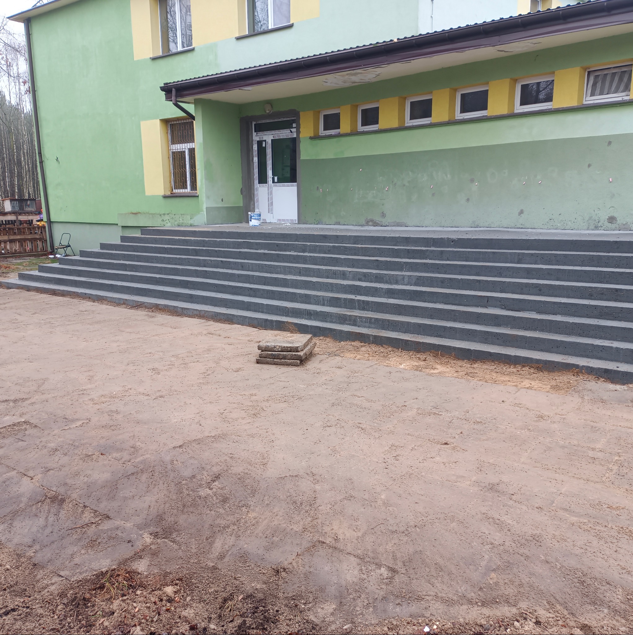 Nowe, betonowe schody wejściowe do budynku z zielono-żółtą elewacją. Widoczny fragment elewacji, drzwi wejściowe i teren wokół schodów.