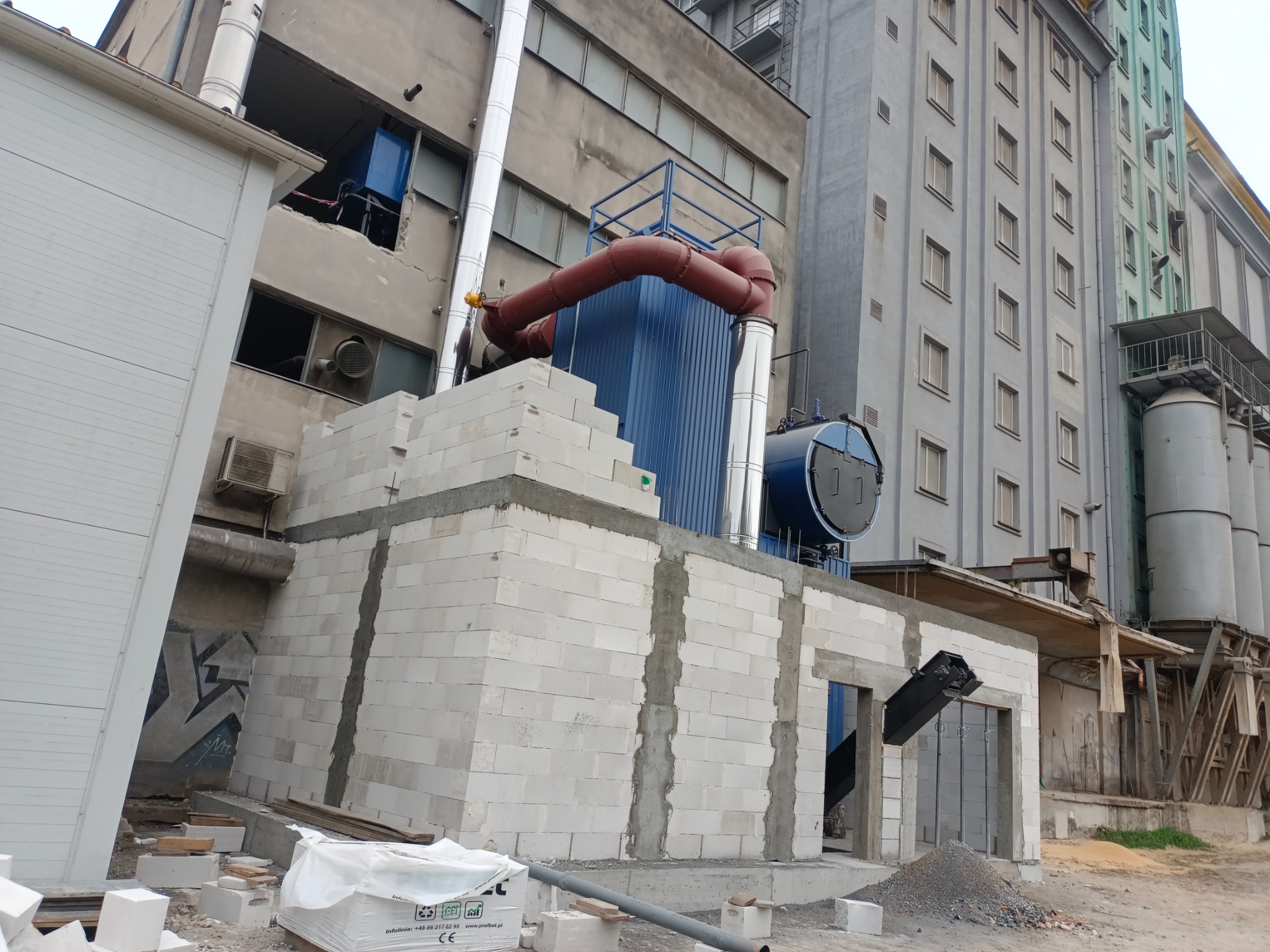 Budowa przyłącza do budynku przemysłowego z białych bloczków. Widoczne rury, komin i elementy instalacji. W tle wysoki budynek z oknami i silosy.