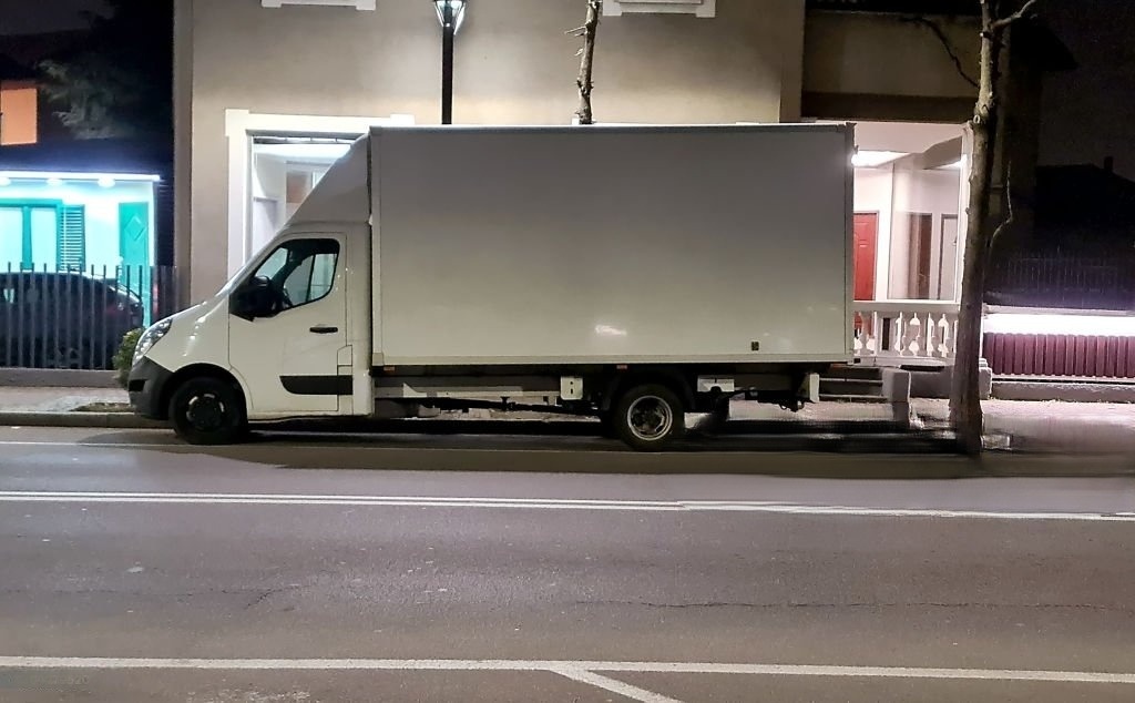 Biały furgon dostawczy zaparkowany nocą przy ulicy, widok z boku, oświetlony światłem ulicznym i reflektorami pobliskich budynków.