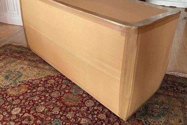 Zapakowany w karton i zabezpieczony taśmą duży przedmiot, prawdopodobnie antyk, leżący na wzorzystym dywanie w pomieszczeniu z parkietem. Przygotowanie do transportu.