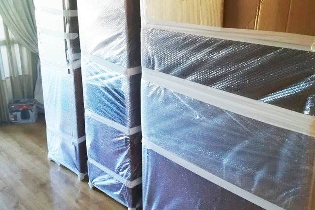 Zabezpieczone meble folią bąbelkową i taśmą, gotowe do transportu. Widoczne trzy zapakowane szafki, stojące na drewnianej podłodze w jasnym pomieszczeniu.