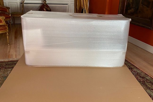 Spakowany antyk w folii bąbelkowej, zabezpieczony kartonem na podłodze, gotowy do transportu. W tle widoczny fragment klasycznego wnętrza.