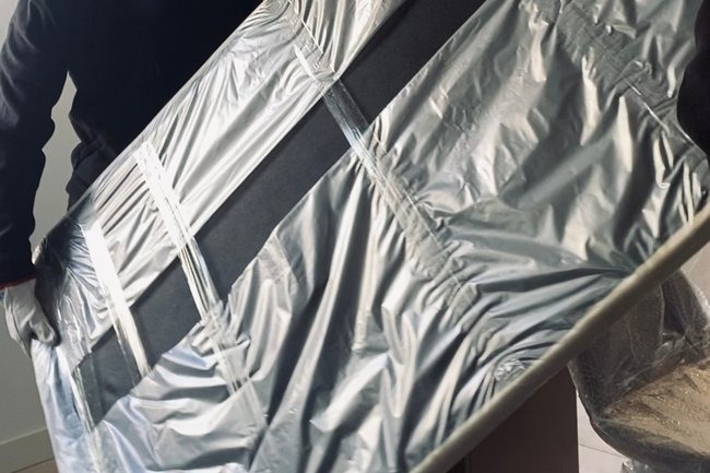 Osoba w rękawicach transportuje zabezpieczony folią i taśmą duży, płaski przedmiot; prawdopodobnie mebel lub obraz, przygotowany do przewozu.