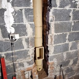 SYSTEMY KOMINOWE Szymon Stokowy - Montaż pionowego przewodu kominowego z otworem rewizyjnym w bloku z szarej cegły. Widoczne narzędzia i poziomice w pomieszczeniu.