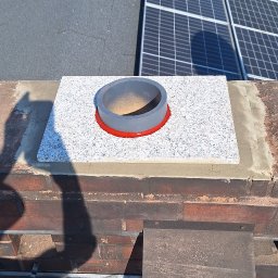 SYSTEMY KOMINOWE Szymon Stokowy - Wykończony komin z szarym wkładem i granitową płytą na dachu, w tle panele słoneczne i budynek. Widoczny cień osoby fotografującej.