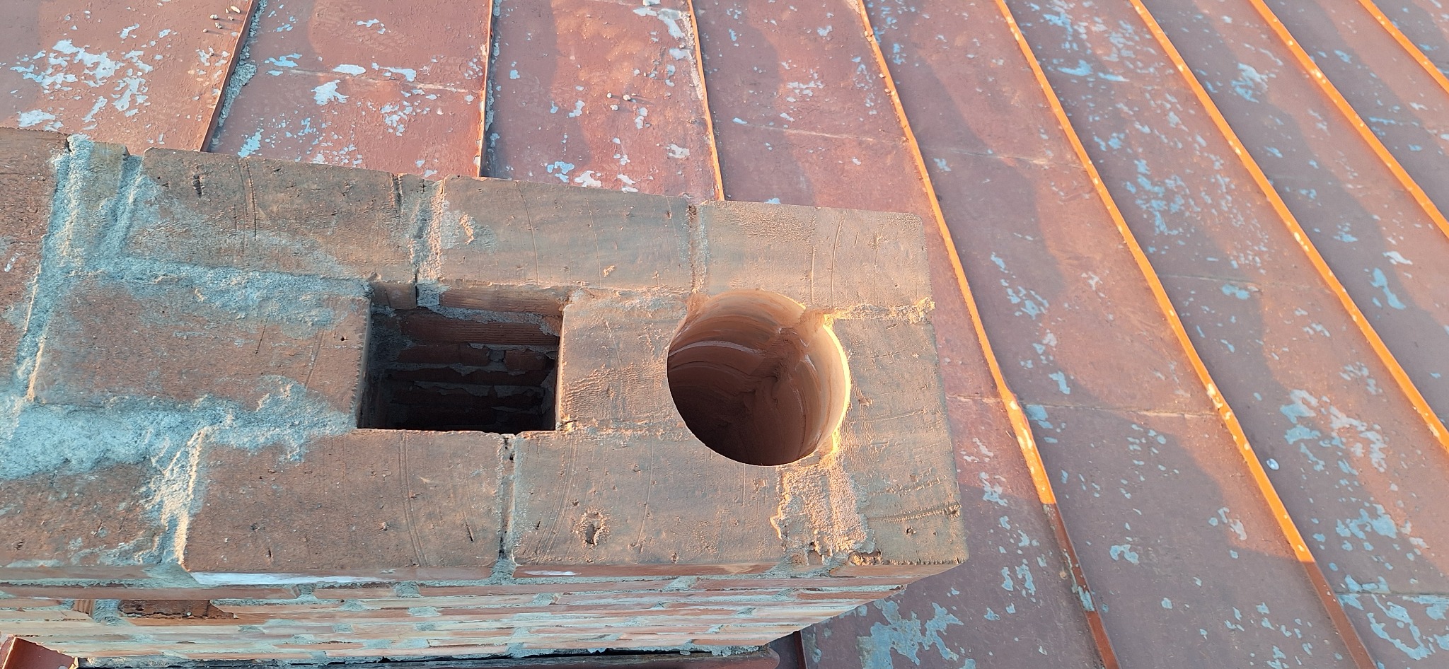 Murowany komin z dwoma otworami na dachu z blachy, oświetlony ciepłym światłem. Widok z góry na konstrukcję komina.