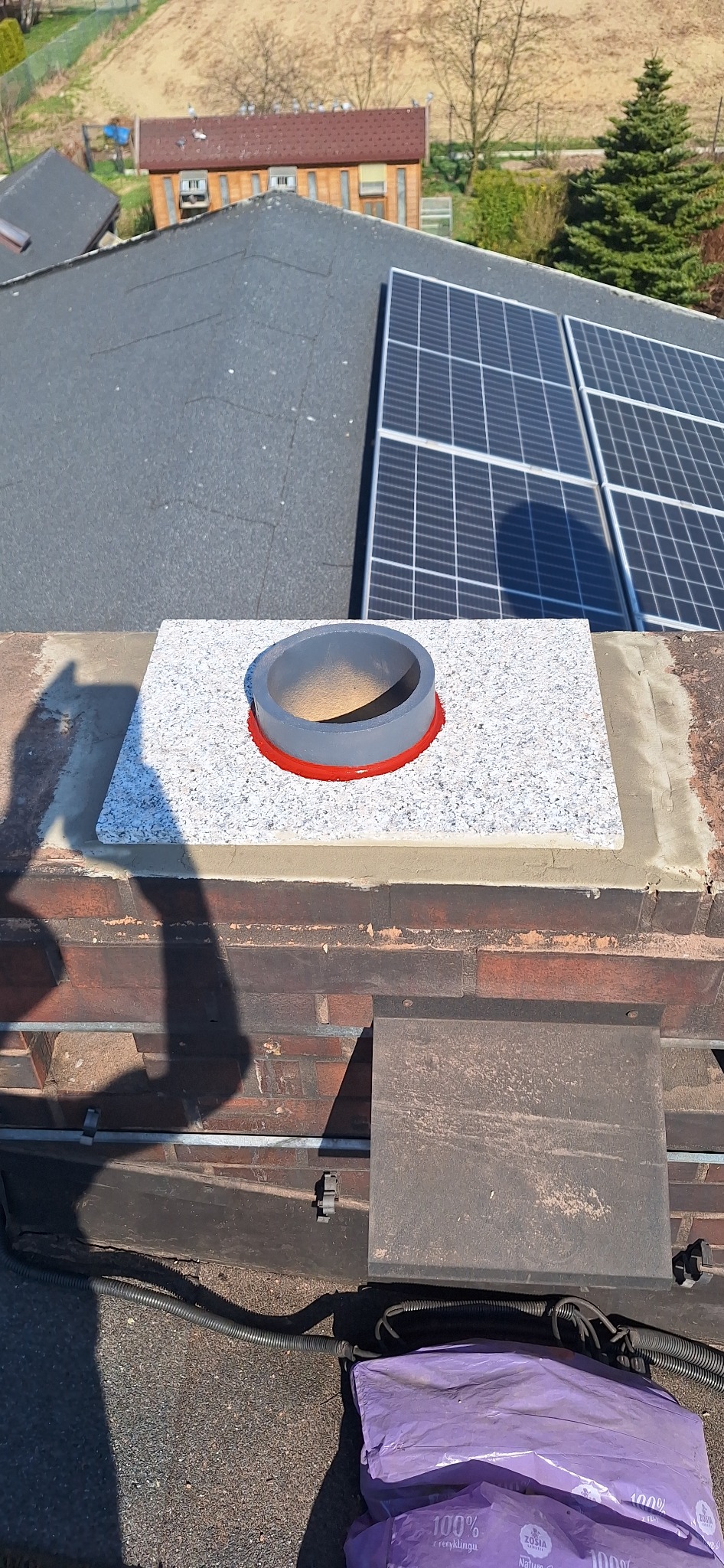 Wykończony komin z szarym wkładem i granitową płytą na dachu, w tle panele słoneczne i budynek. Widoczny cień osoby fotografującej.