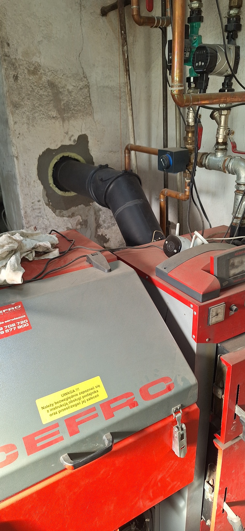 Podłączenie rury spalinowej do ściany, widoczny piec CO Defro z instrukcją obsługi. Instalacja grzewcza z miedzianych rur. Szare tło ściany.