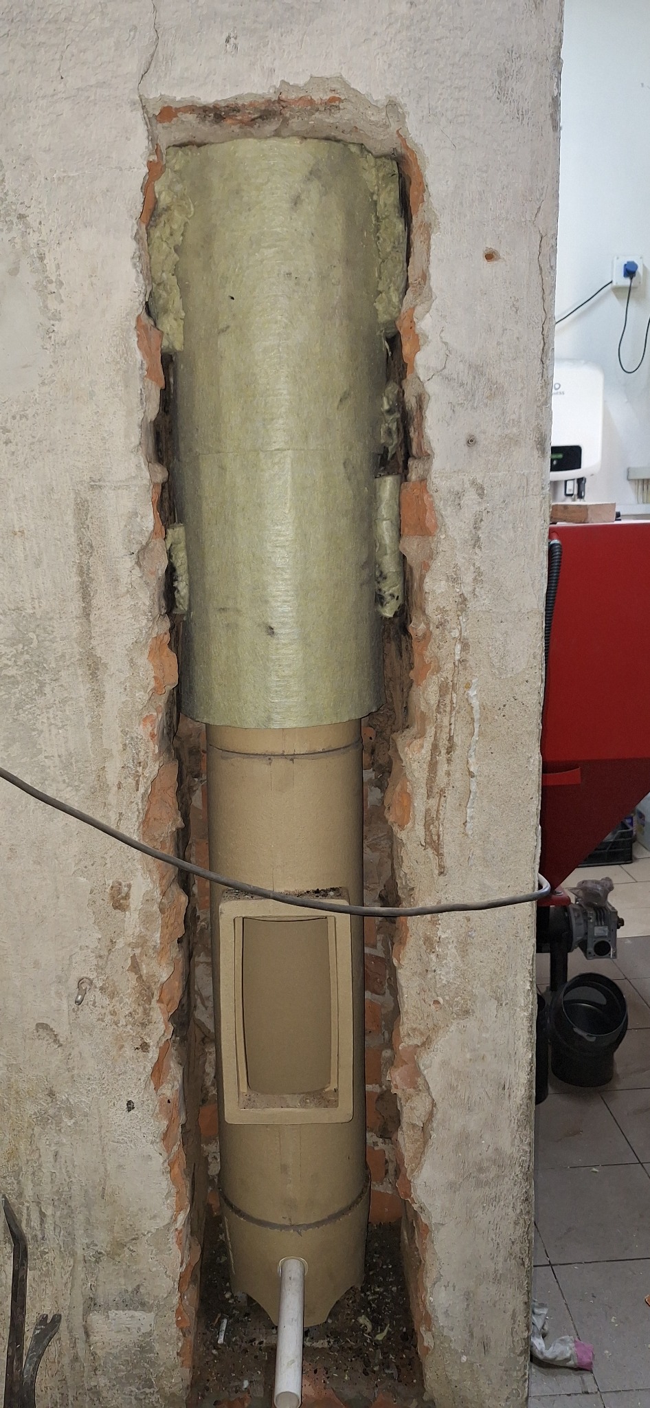 Ocieplony wkład kominowy z otworem rewizyjnym, zamontowany w wykuciu ściany z widocznymi cegłami. Element kotłowni w tle.