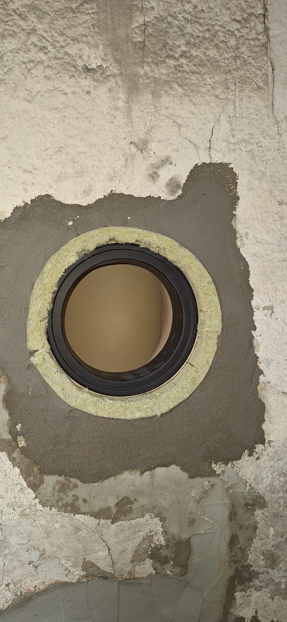 Okrągły wlot kominowy z izolacją termiczną, osadzony w surowej ścianie. Widoczne ślady zaprawy i nierówności tynku.