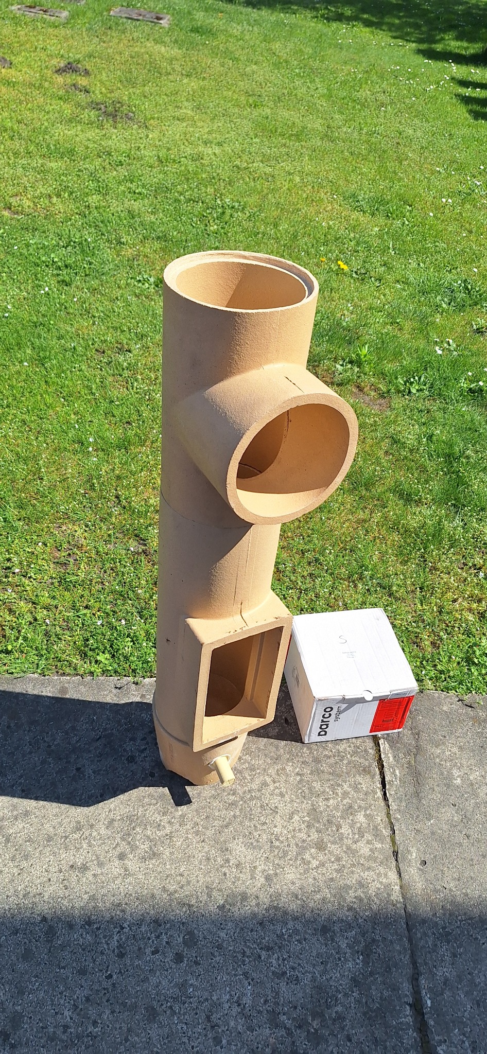 Beżowy prefabrykowany komin ceramiczny z otworem bocznym i wyczystką, obok kartonowe opakowanie na tle trawnika.