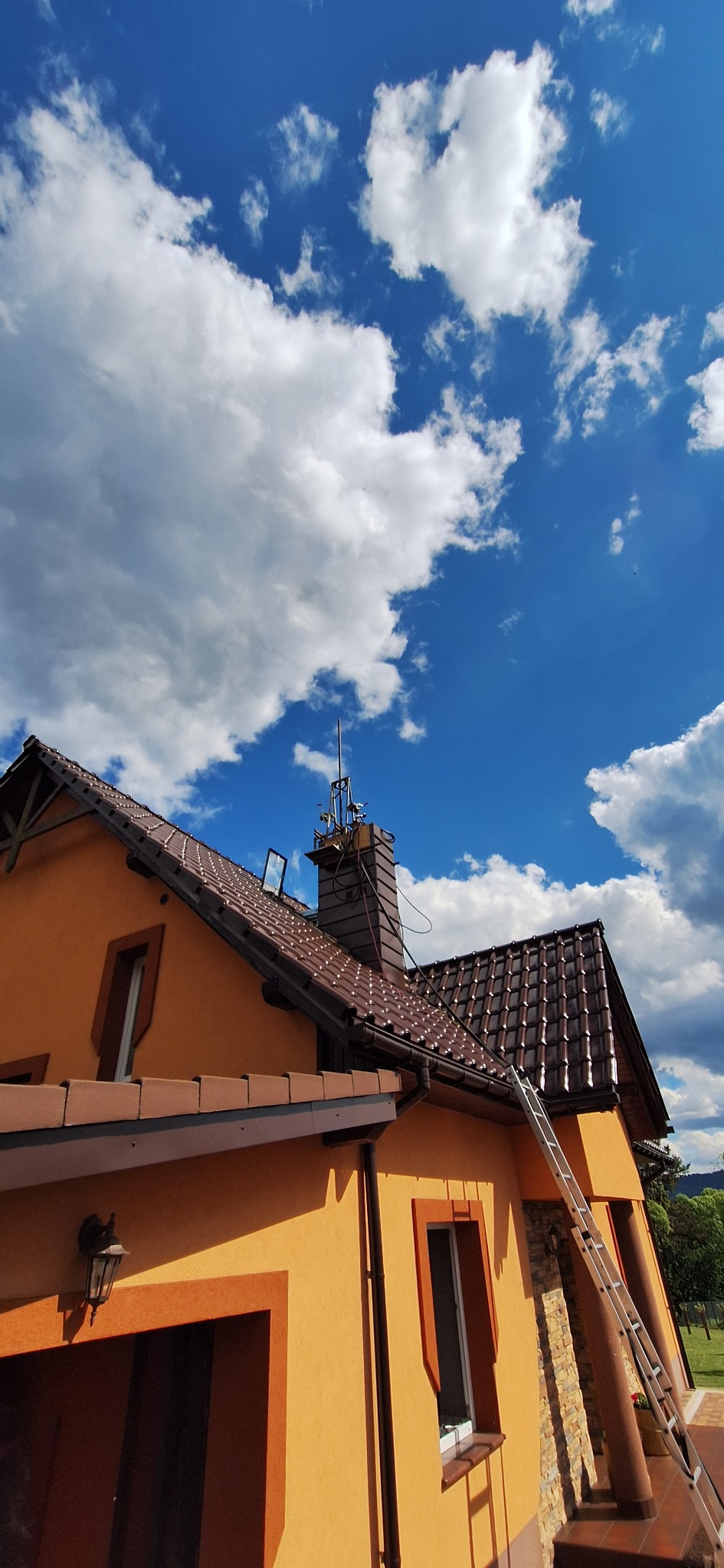 Żółty dom z brązowym dachem i kominem, drabina oparta o ścianę, błękitne niebo z chmurami w tle. Widok z dołu.