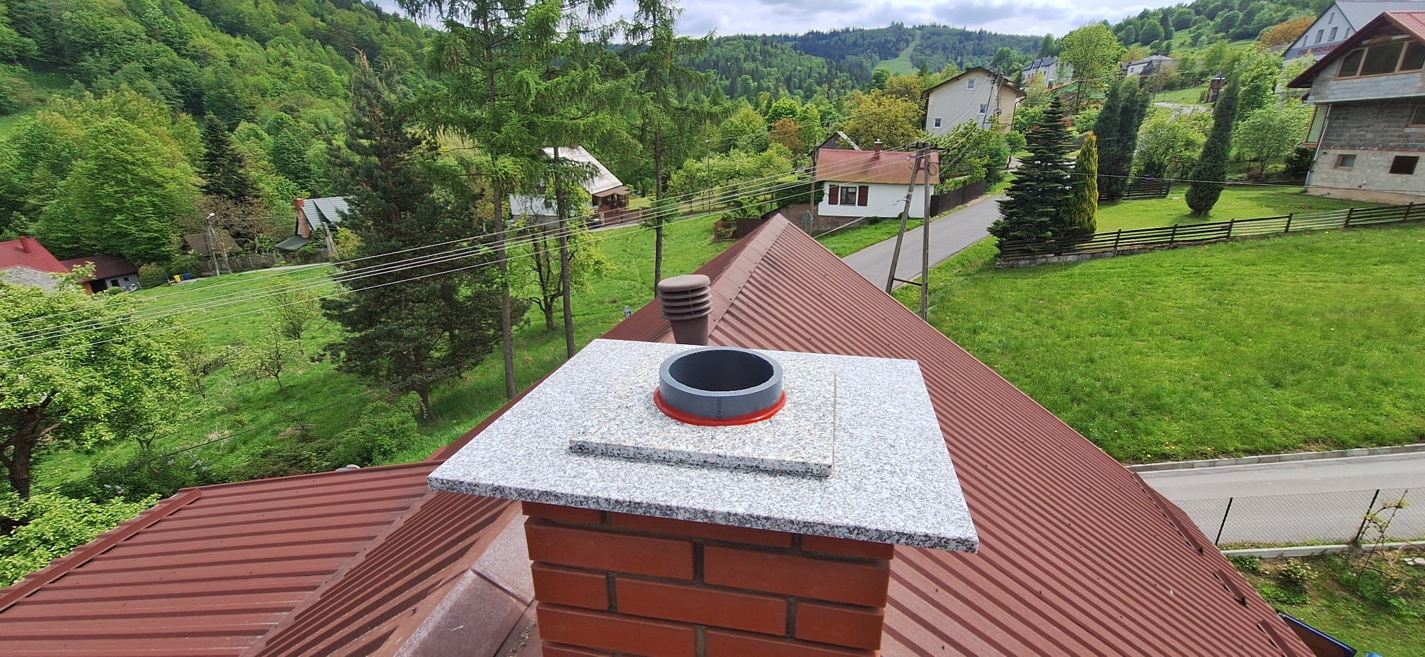 Wykończony komin z cegły, z szarą płytą i elementami instalacji. Tło to zielone wzgórza i domy. Widok z góry na brązowy dach.