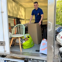 BOXERSI GROUP SPÓŁKA Z OGRANICZONĄ ODPOWIEDZIALNOŚCIĄ - Uśmiechnięty mężczyzna w koszulce z logo firmy przeprowadzkowej wewnątrz busa załadowanego meblami i kartonami, gotowego do transportu. Widoczny fotel i torby.