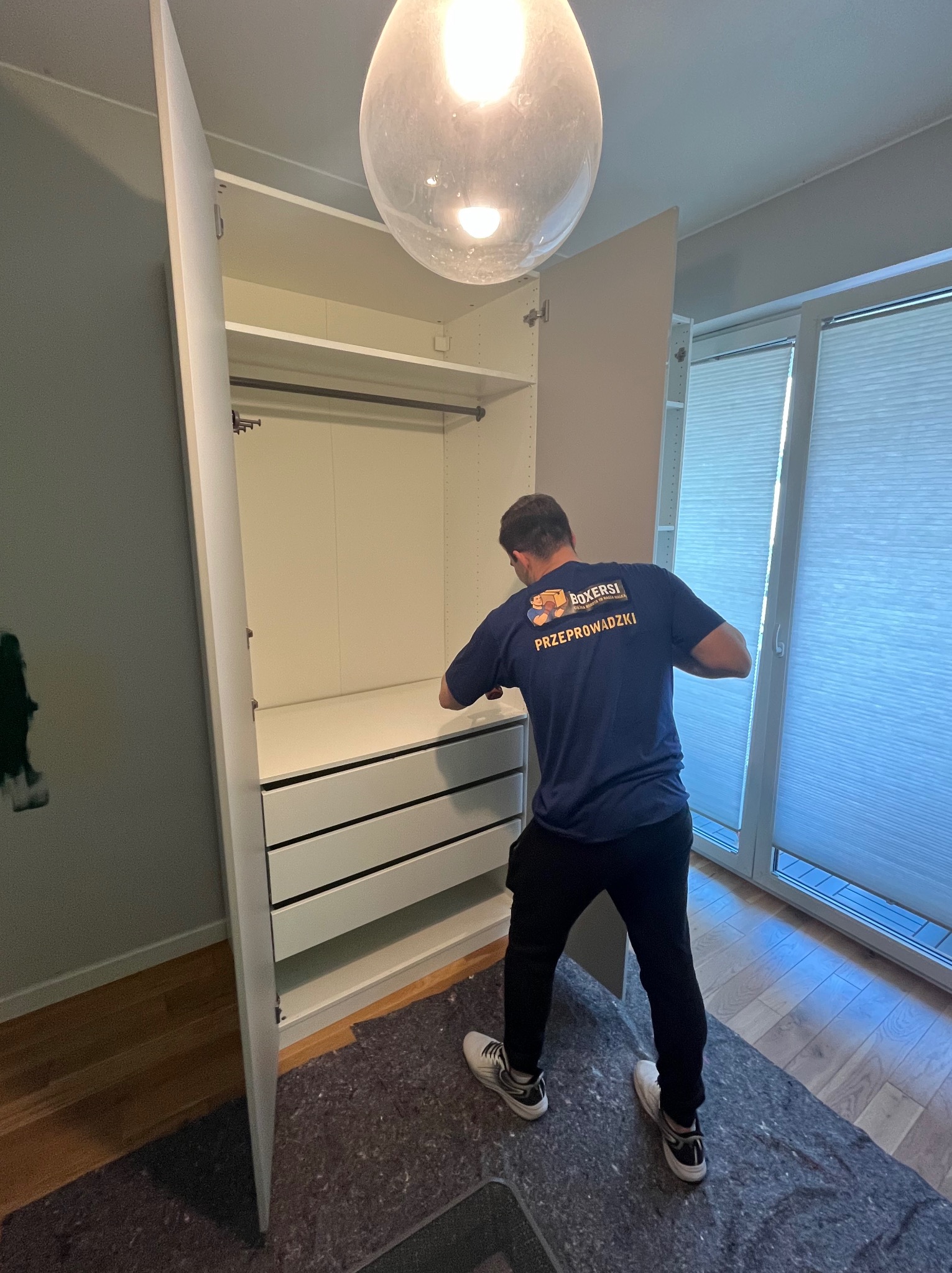 Mężczyzna w niebieskiej koszulce z logo firmy przeprowadzkowej montuje białą szafę z szufladami w jasnym wnętrzu. Widoczne otwarte drzwi szafy i rolety okienne.