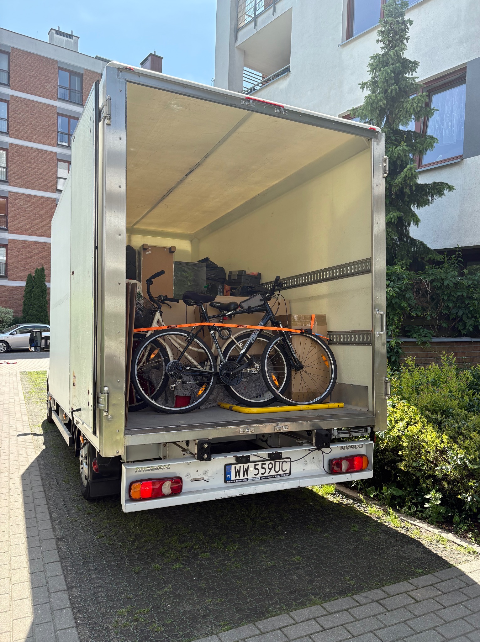 Otwarta paka busa transportowego z dwoma rowerami i kartonami w środku. Rejestracja WW 559UC. Tło: budynek mieszkalny i zieleń. Słoneczny dzień.