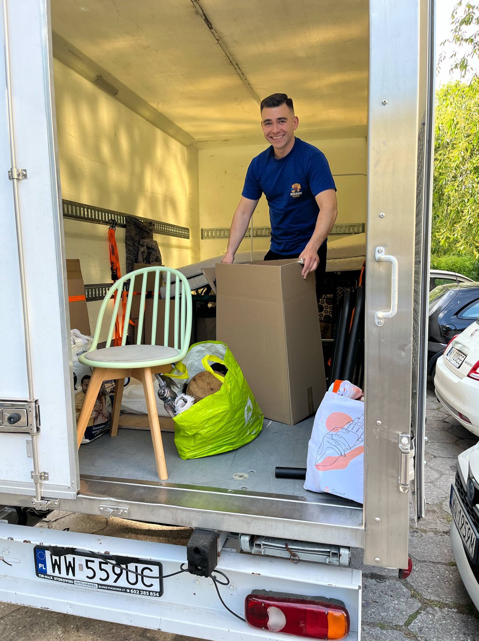 Uśmiechnięty mężczyzna w koszulce z logo firmy przeprowadzkowej wewnątrz busa załadowanego meblami i kartonami, gotowego do transportu. Widoczny fotel i torby.