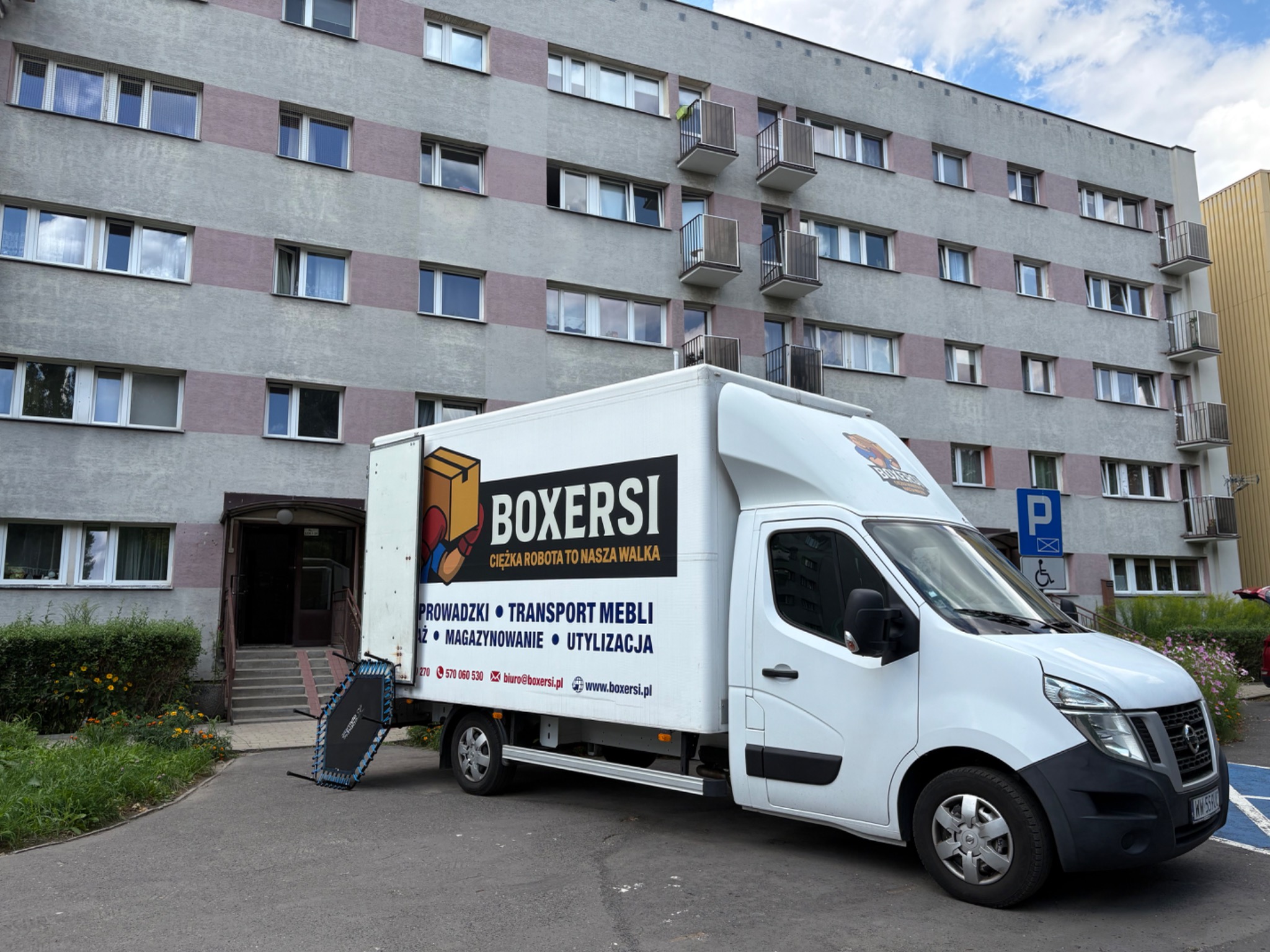Biała furgonetka firmy przeprowadzkowej Boxersi z trampoliną obok wejścia do bloku. Usługi: przeprowadzki, transport mebli, magazynowanie, utylizacja.