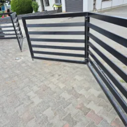 Nowoczesna brama wjazdowa, czarna, poziome profile, częściowo otwarta. Widoczny fragment posesji i kostka brukowa na podjeździe. Brama dwuskrzydłowa.