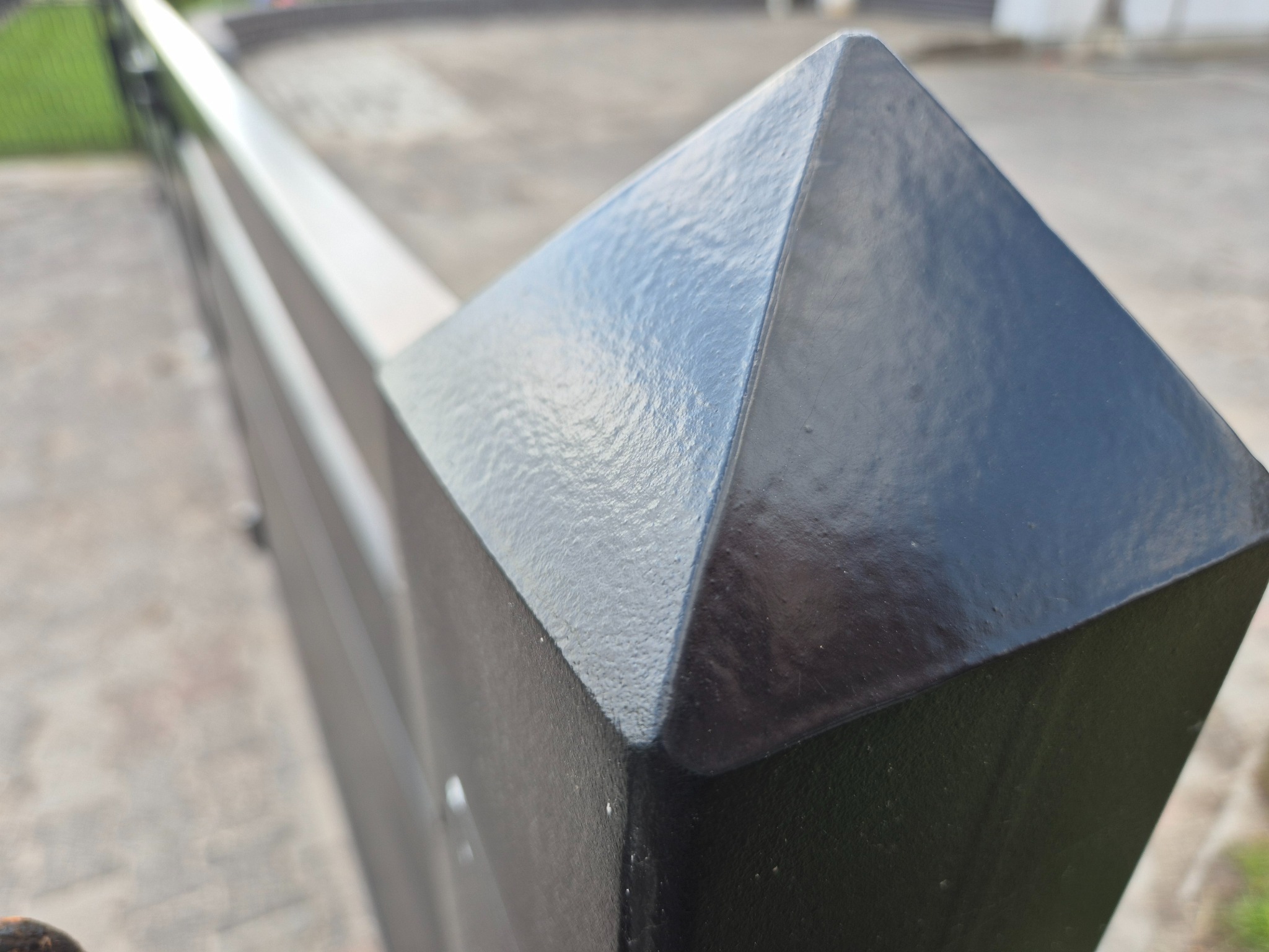 Detal czarnego, metalowego słupka ogrodzeniowego z piramidalnym zakończeniem. Widoczne łączenia spawów i faktura malowanej powierzchni, fragment chodnika w tle.