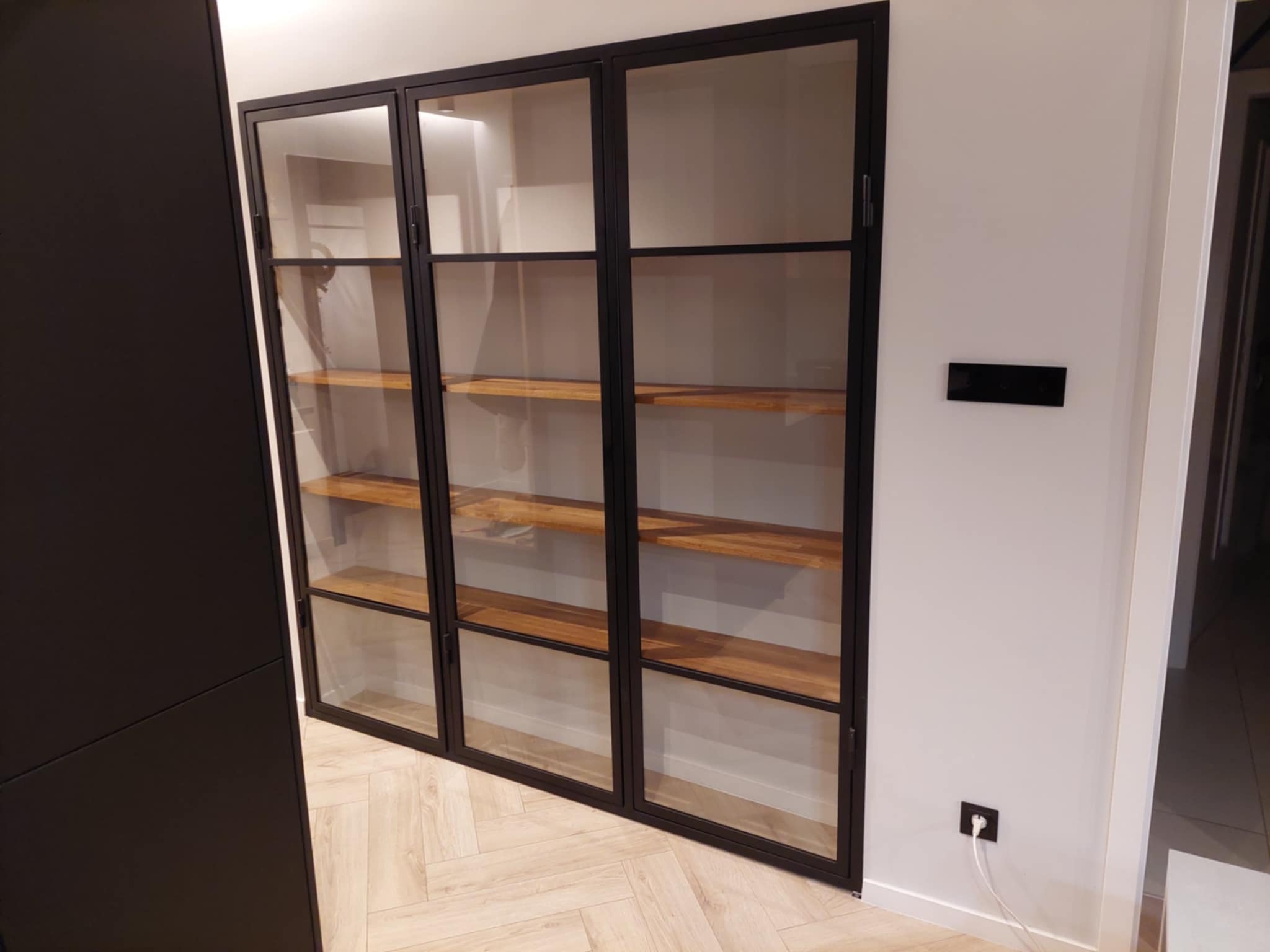 Nowoczesna, przeszklona szafa z czarnymi metalowymi ramami i drewnianymi półkami, wkomponowana we wnętrze z jasną podłogą i białymi ścianami.