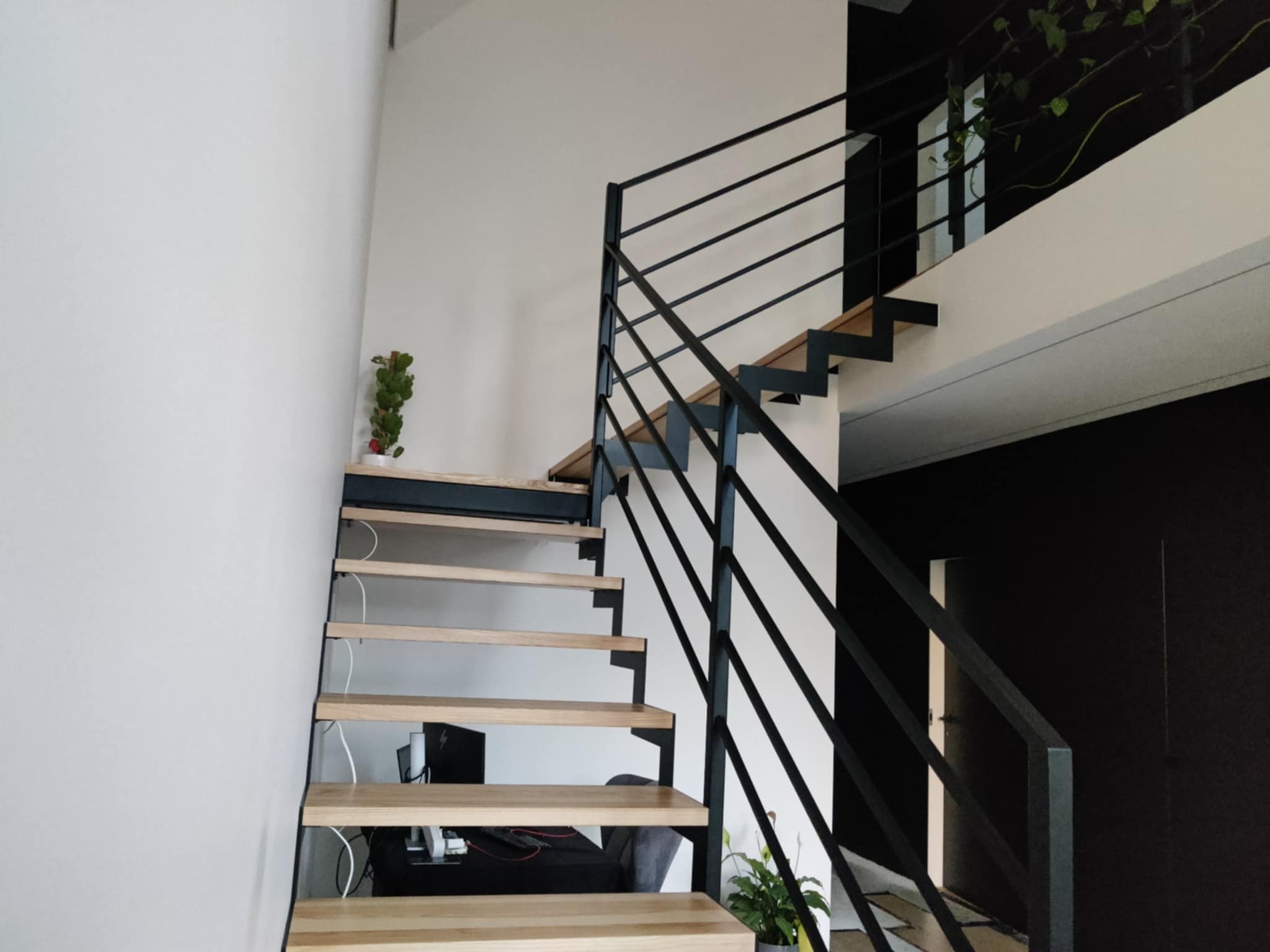 Nowoczesne schody z drewnianymi stopniami i czarną metalową balustradą w minimalistycznym wnętrzu. Widok z dołu, podkreślający geometrię konstrukcji.