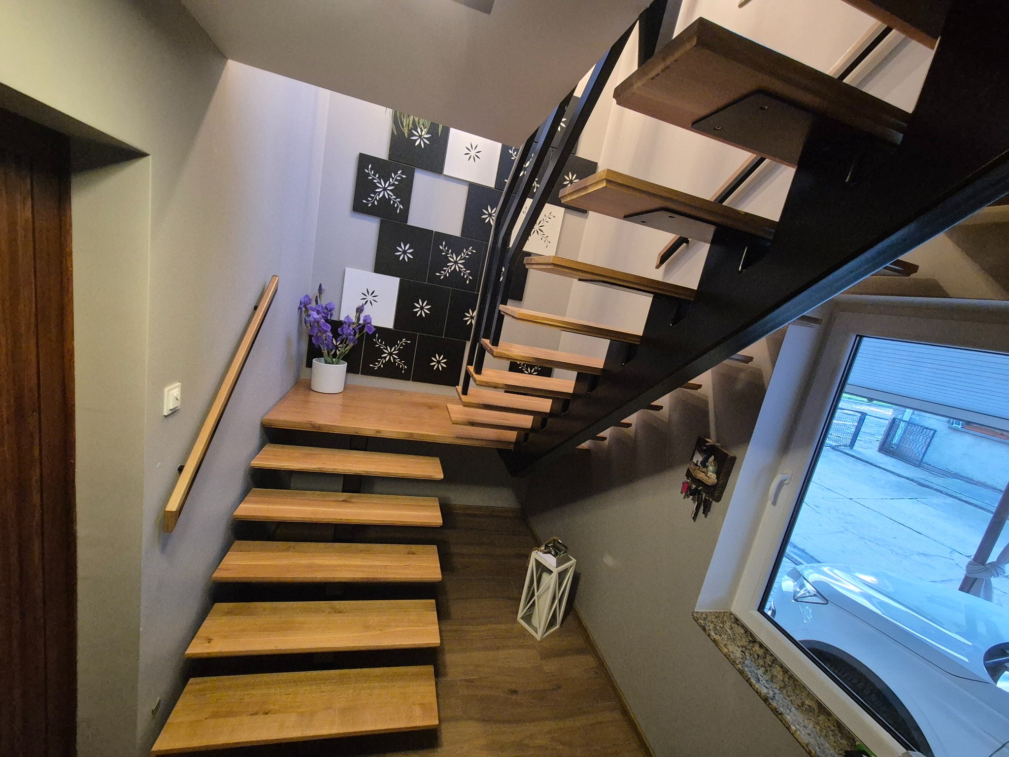 Nowoczesne schody drewniane z metalową konstrukcją w jasnym wnętrzu. Na ścianie dekoracyjne płytki i drewniana poręcz. Widok z góry na stopnie i biały lampion.