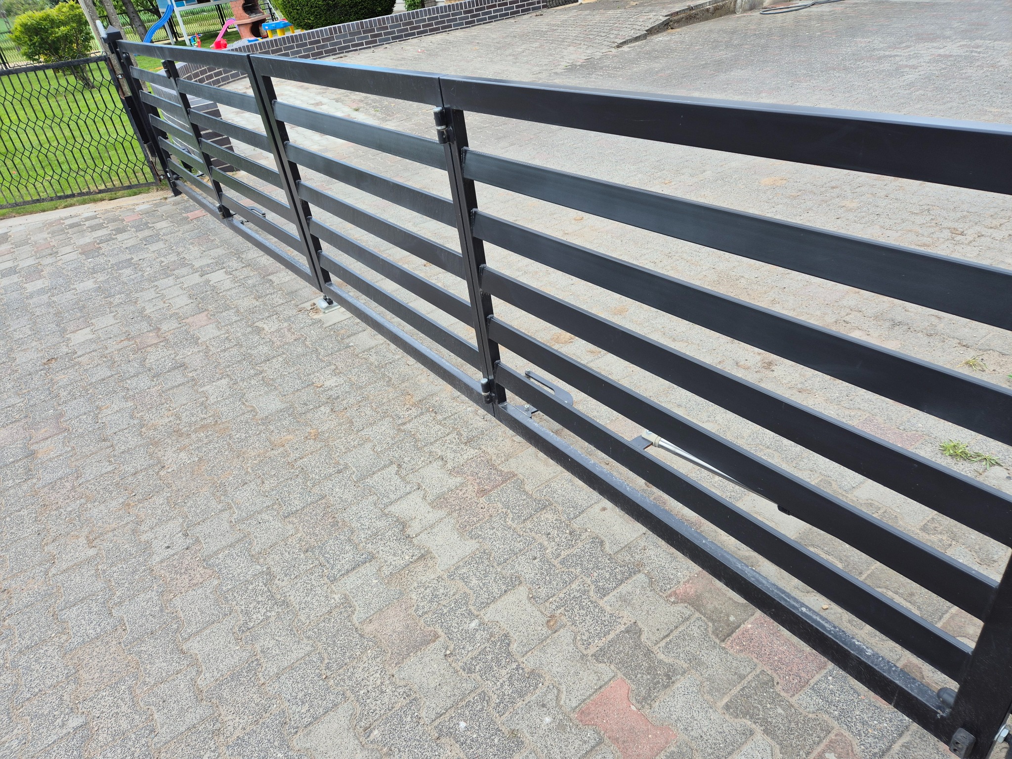 Nowoczesna brama wjazdowa z poziomych, grafitowych profili stalowych, zamontowana na posesji wyłożonej kostką brukową. Widoczny mechanizm otwierania.